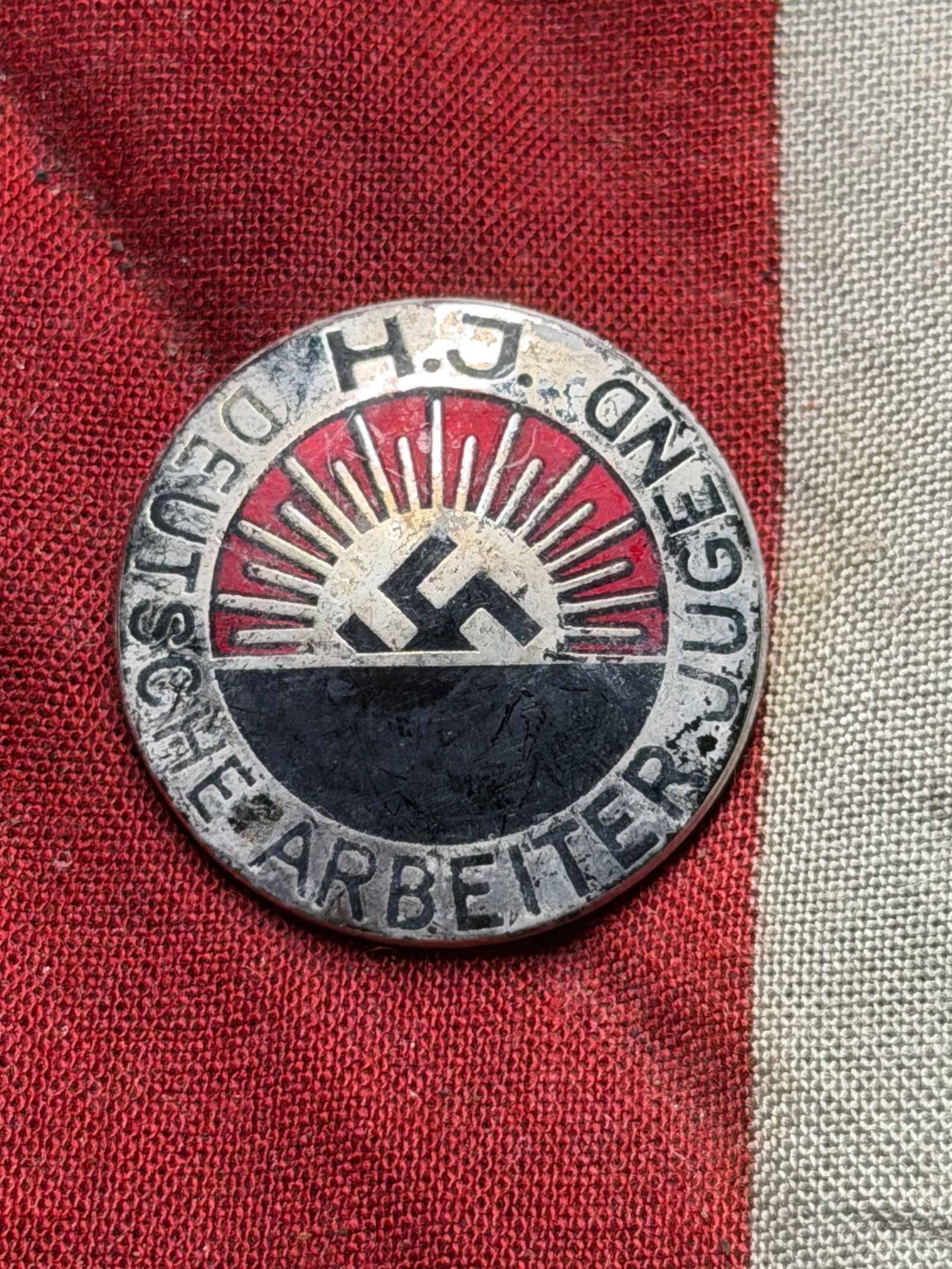 WW2 German Hitler Youth Membership Badge (H.J. – Deutsche Arbeiterjugend) By RZM M1/129: This circular enameled metal badge bears the inscription “H.J. Deutsche Arbeiterjugend” around a central motif incorporating a stylized rising sun design. The reverse features a pin-back attac