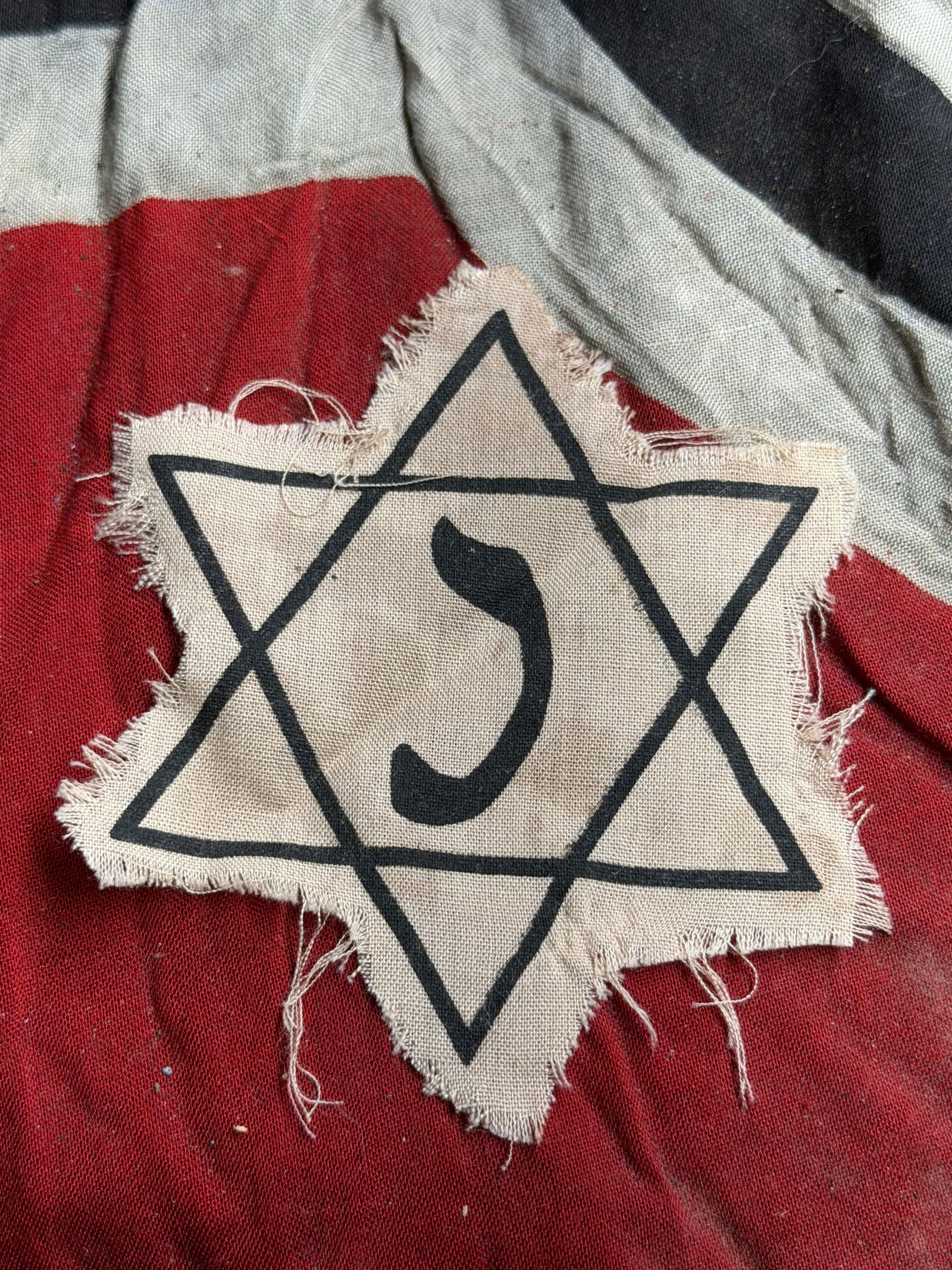 Jewish Holocaust Identification Badge (Star of David Armband Fragment), World War II Era: This cloth badge, made of beige fabric and printed with a black Star of David enclosing a Hebrew letter, was part of the system of identification imposed on Jewish civilians during the Nazi occupation