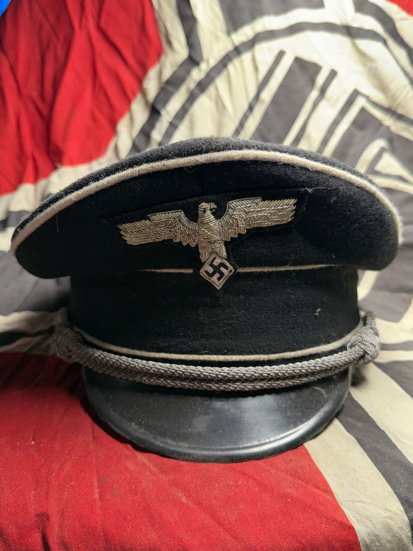 WWII German Officer’s Visor Cap – Black Wool with Silver Piping and Bullion Insignia: This listing features a World War II-era German officer’s visor cap, constructed of black wool with silver piping and adorned with hand-embroidered bullion insignia on the front. The cap has the tra
