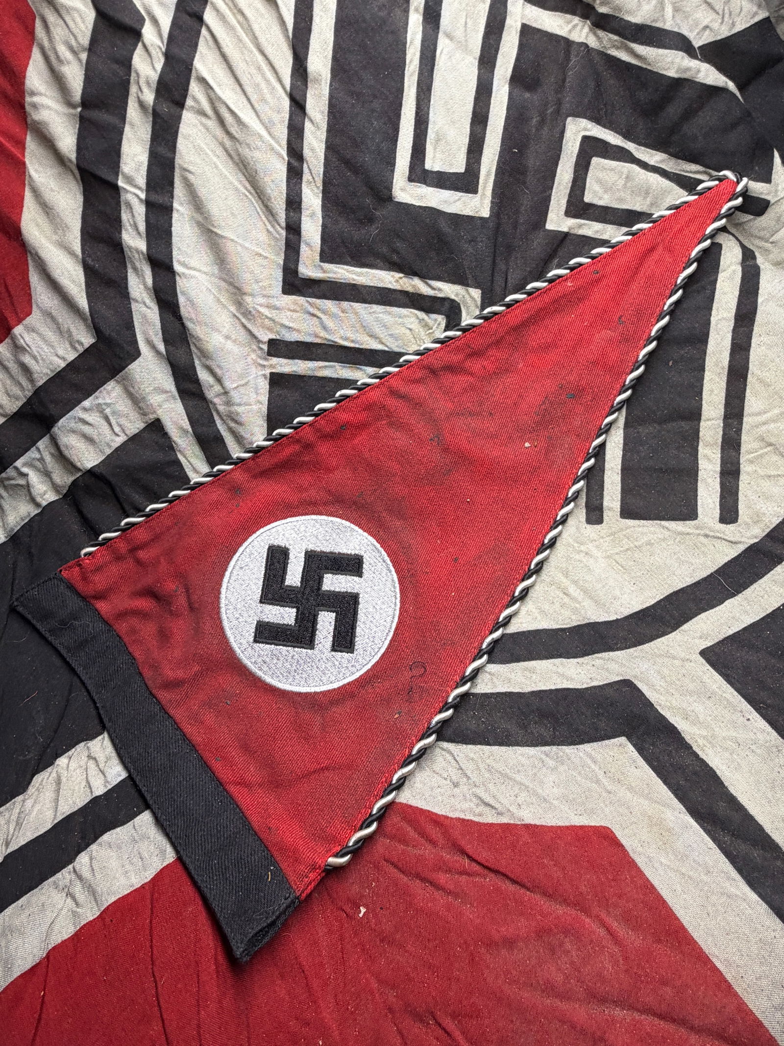 WW2 German NSDAP National Socialist Pennant, 1930s–1940s: This triangular fabric pennant features a red field with a central white circle and black hooked cross symbol, edged with twisted black-and-white cord. Measuring approximately 12 inches in length, the