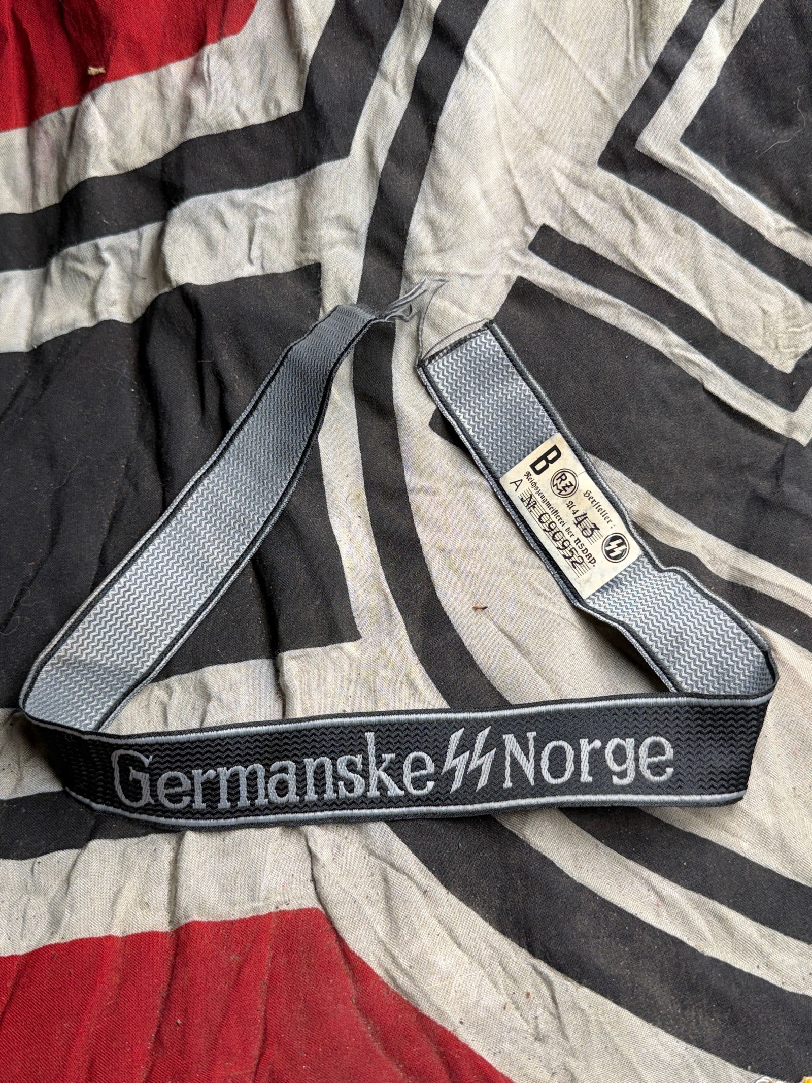 WW2 German Germanske Norge Cuff Title With RZM Paper Tag: WW2 German Germanske Norge Cuff Title With RZM Paper Tag. This black and silver-grey woven cuff title bears the text “Germanske Norge” embroidered in silver-grey thread, bordered by chevron-pa