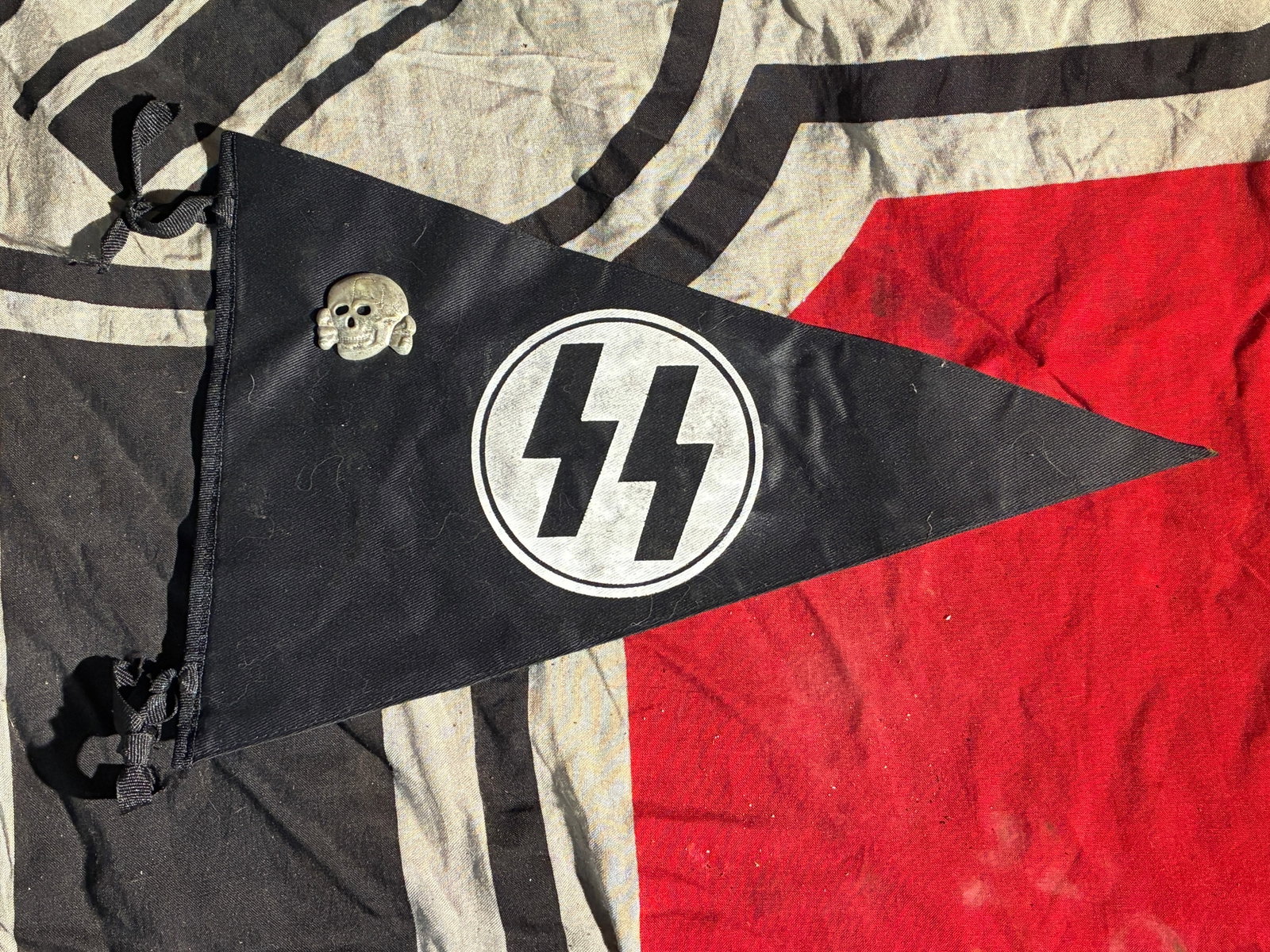 WW2 German SS Pennant With Attached Totenkopf Cap Badge (c. 1930s–1945): This triangular black cloth pennant features the insignia of the Schutzstaffel (SS), one of Nazi Germany’s principal organizations responsible for systemic crimes including mass repression and the g