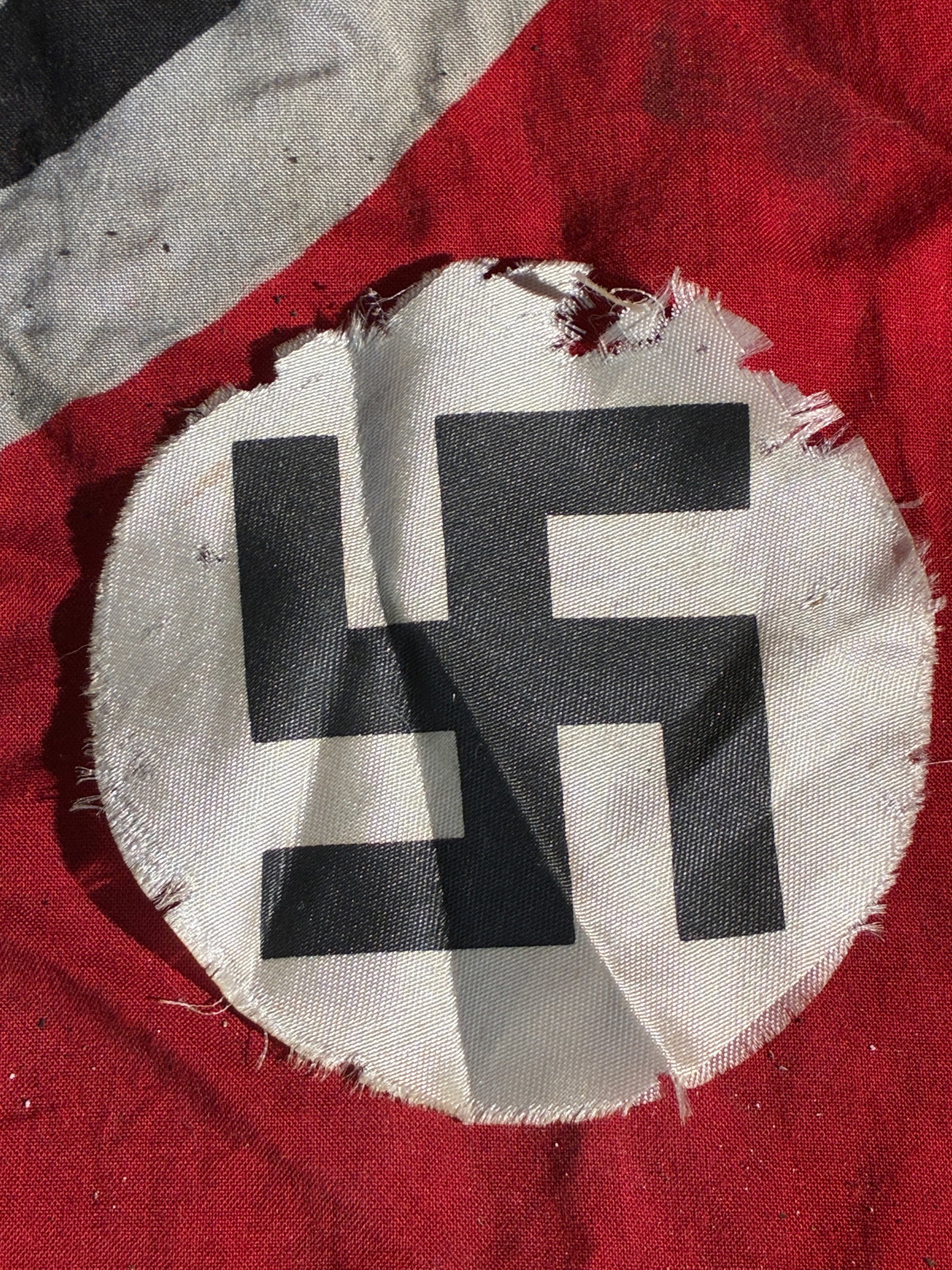 WW2 German Fragment of a Nazi Party Flag Roundel (Swastika Emblem), ca. 1930s–1945: This object is a heavily worn and torn circular cloth roundel removed from a Nazi Party flag or banner. The roundel displays a printed black swastika on a white field—one of the central components o