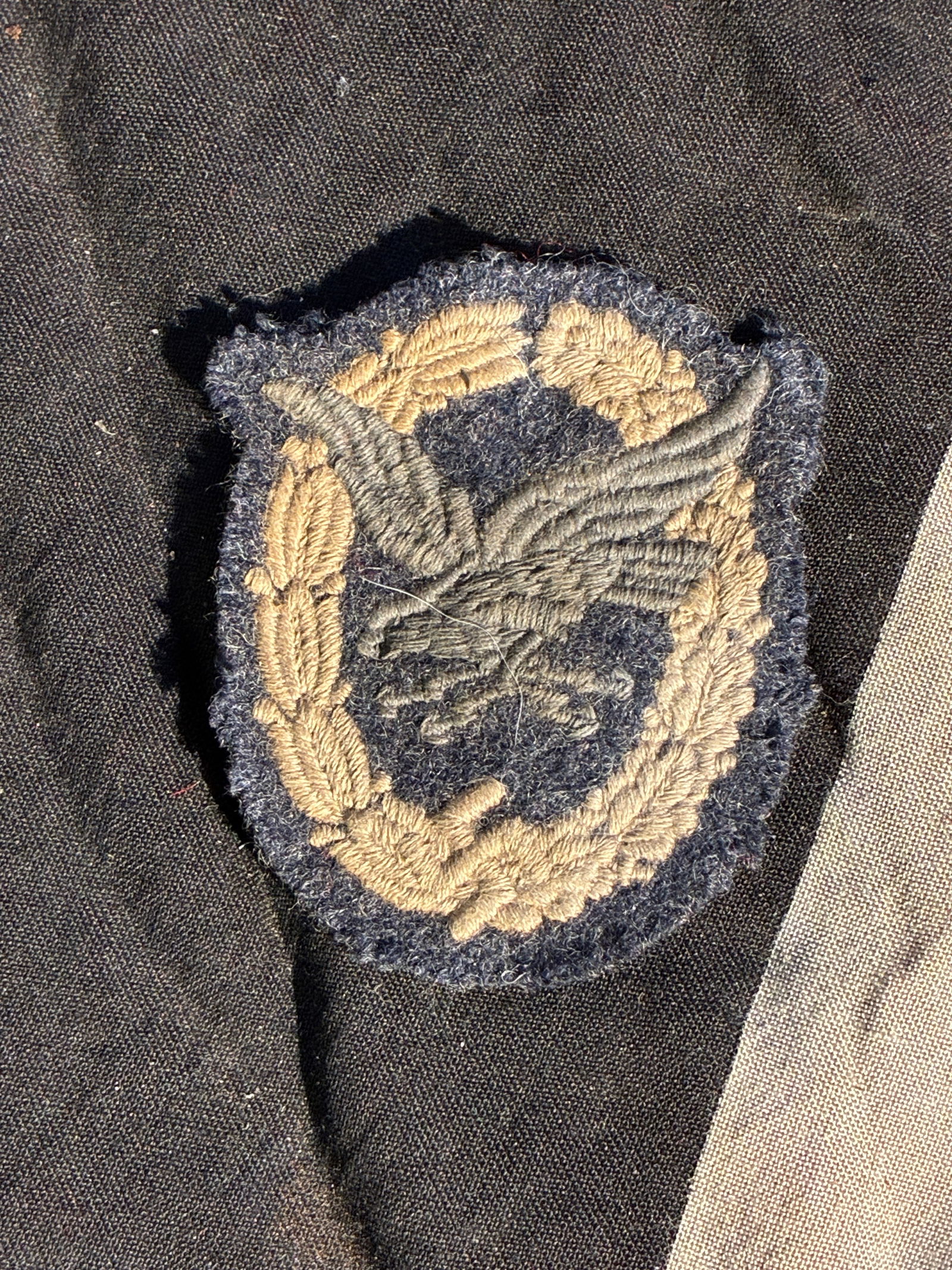 WW2 German Luftwaffe Cloth Pilot’s Badge (Worn Variant), c. 1939–1945: This heavily worn Luftwaffe cloth pilot’s badge was issued to flight personnel of Nazi Germany’s air force during the Second World War. Embroidered in grey and tan thread on a dark blue wool backi