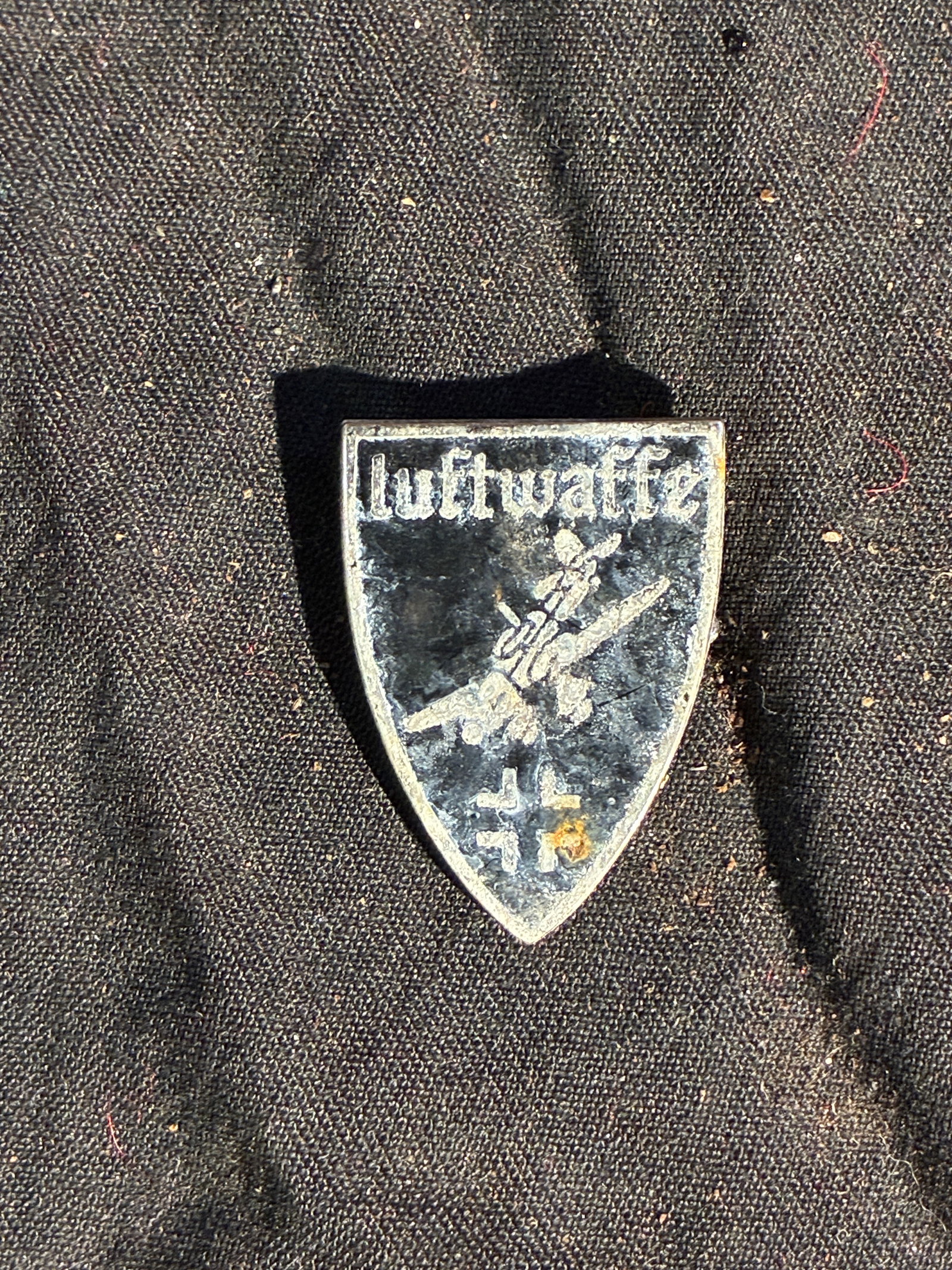 WW2 German Air Force – WWII Era: This small, shield-shaped souvenir pin features the word “Luftwaffe” above a stylized depiction of an aircraft in flight, accompanied by two Balkenkreuz-style crosses. Produced as a privately 