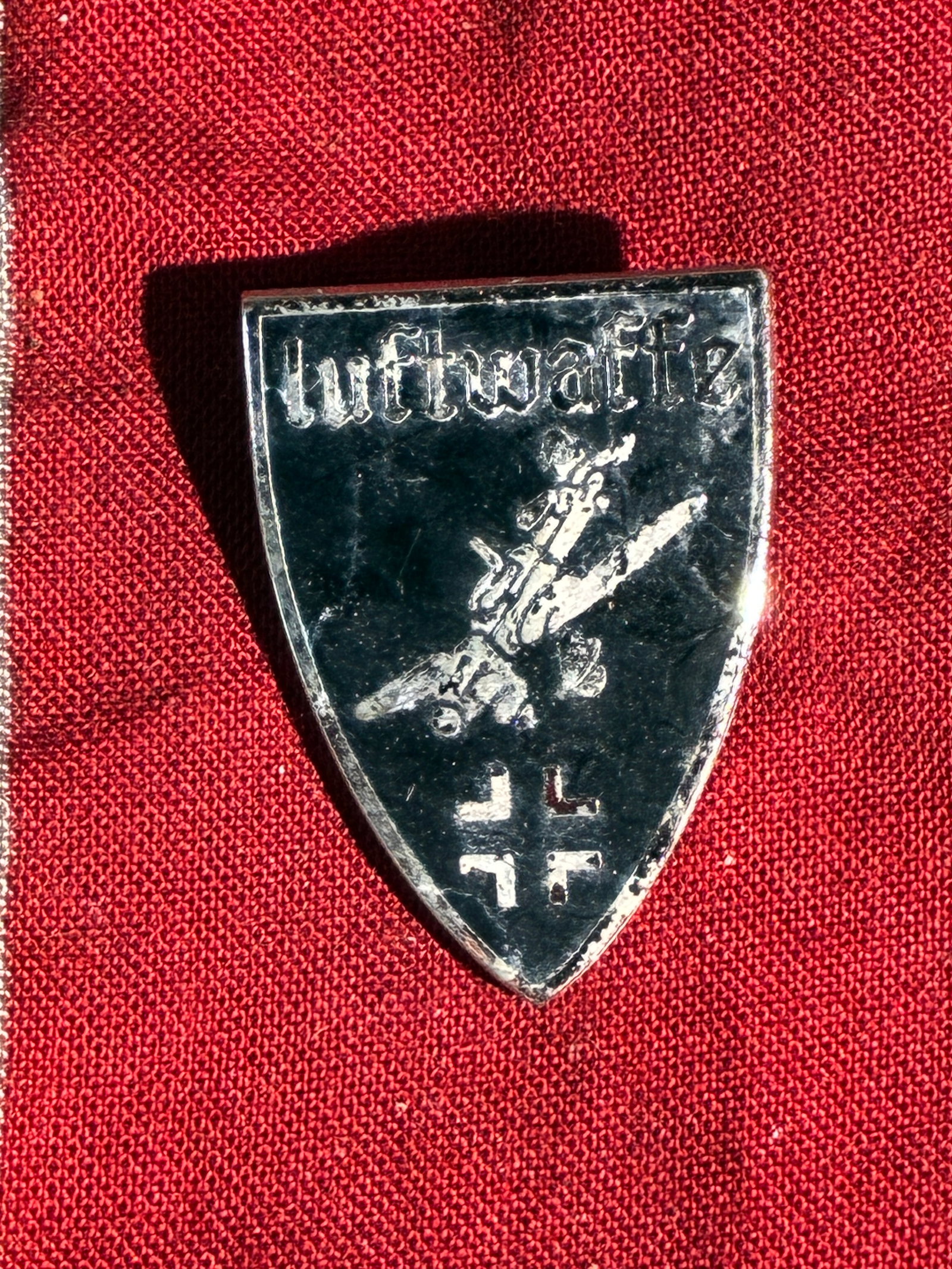 WW2 German Luftwaffe Commemorative Shield Badge (Unofficial / Souvenir Type), Germany, c.: This small shield-shaped badge features the word “Luftwaffe” above a stylized diving aircraft and a pair of swastika motifs, stamped into a bright metal surface. Produced outside the official 