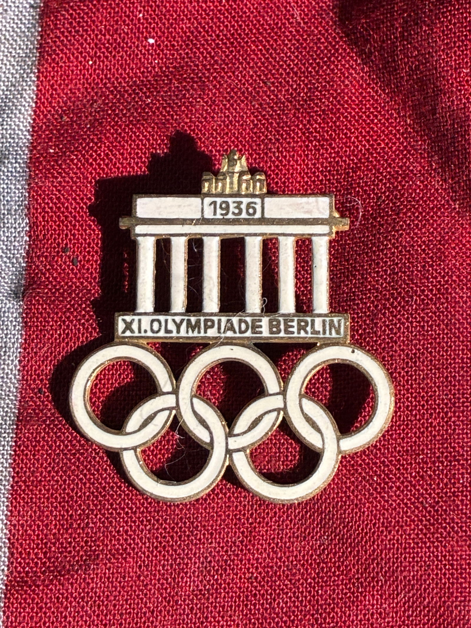 WW2 German Commemorative Pin from the 1936 Berlin Olympic Games: This small enameled pin was produced as a souvenir for the XI Olympiad, held in Berlin in 1936. The design features the iconic Brandenburg Gate, one of Germany’s most recognizable landmarks, positio