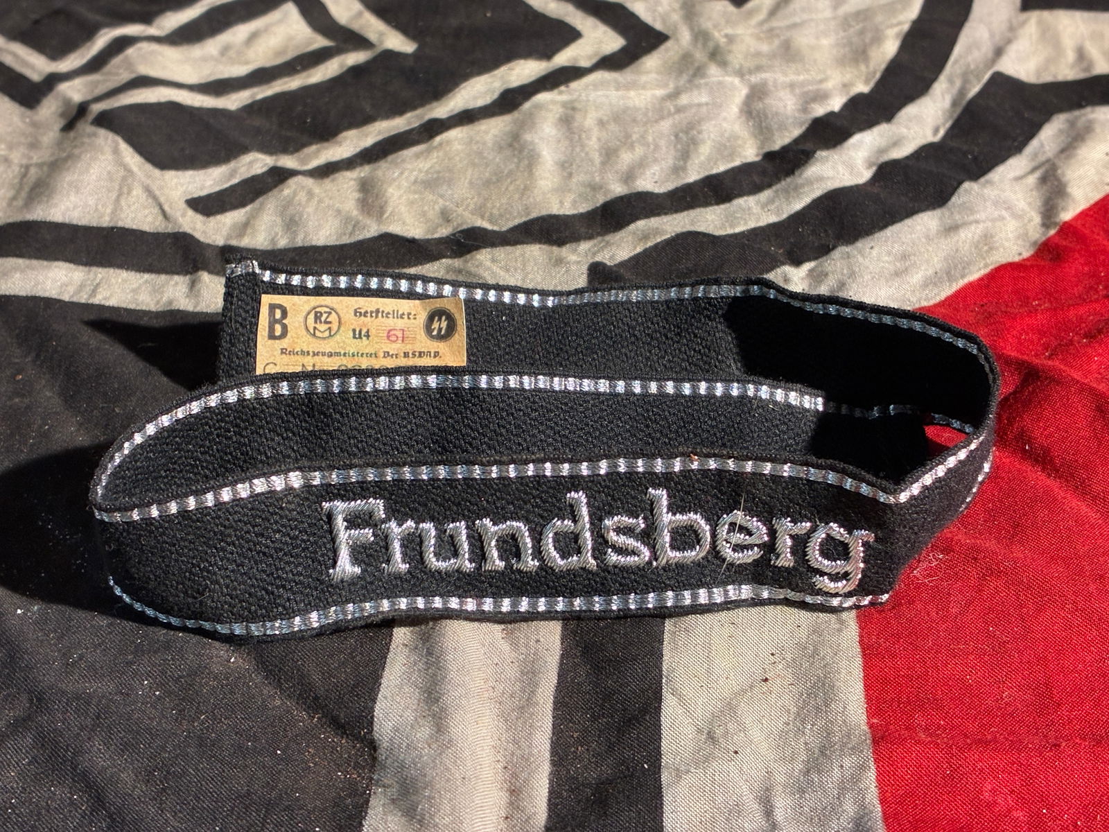 WW2 German Cuff Title of the 10th SS Panzer Division “Frundsberg?? With RZM Paper Tag: This embroidered cuff title belonged to the 10th SS Panzer Division “Frundsberg,” a Waffen-SS armored formation of Nazi Germany active during the Second World War. The black wool band features