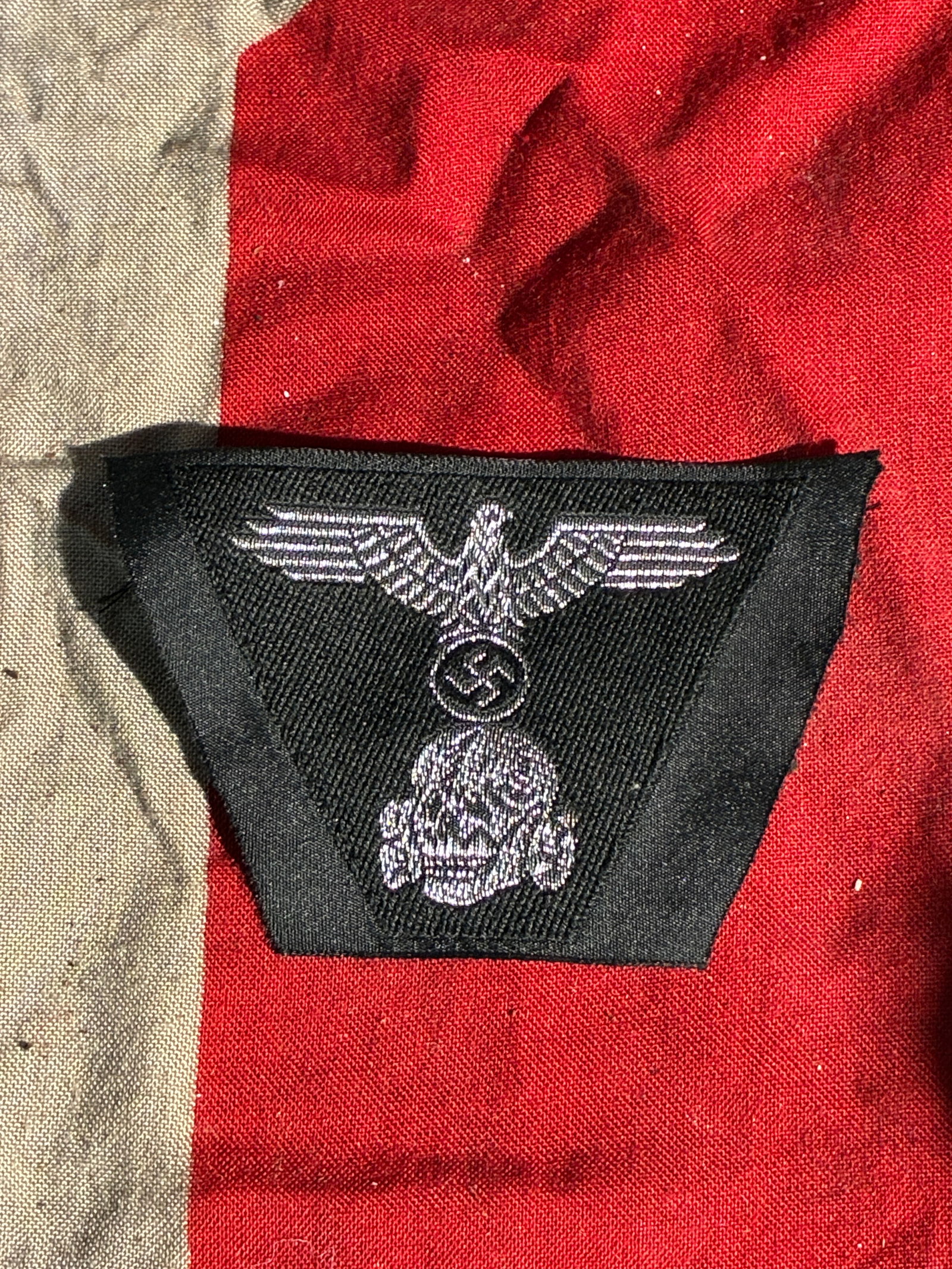 WW2 German Waffen-SS Sleeve Insignia Fragment (Totenkopf & Eagle Motif), c. 1930s–1945: This textile fragment features an embroidered combination of two major symbols used by the Waffen-SS: the SS eagle clutching a swastika and the Totenkopf (skull) emblem. Such insignia were typically w