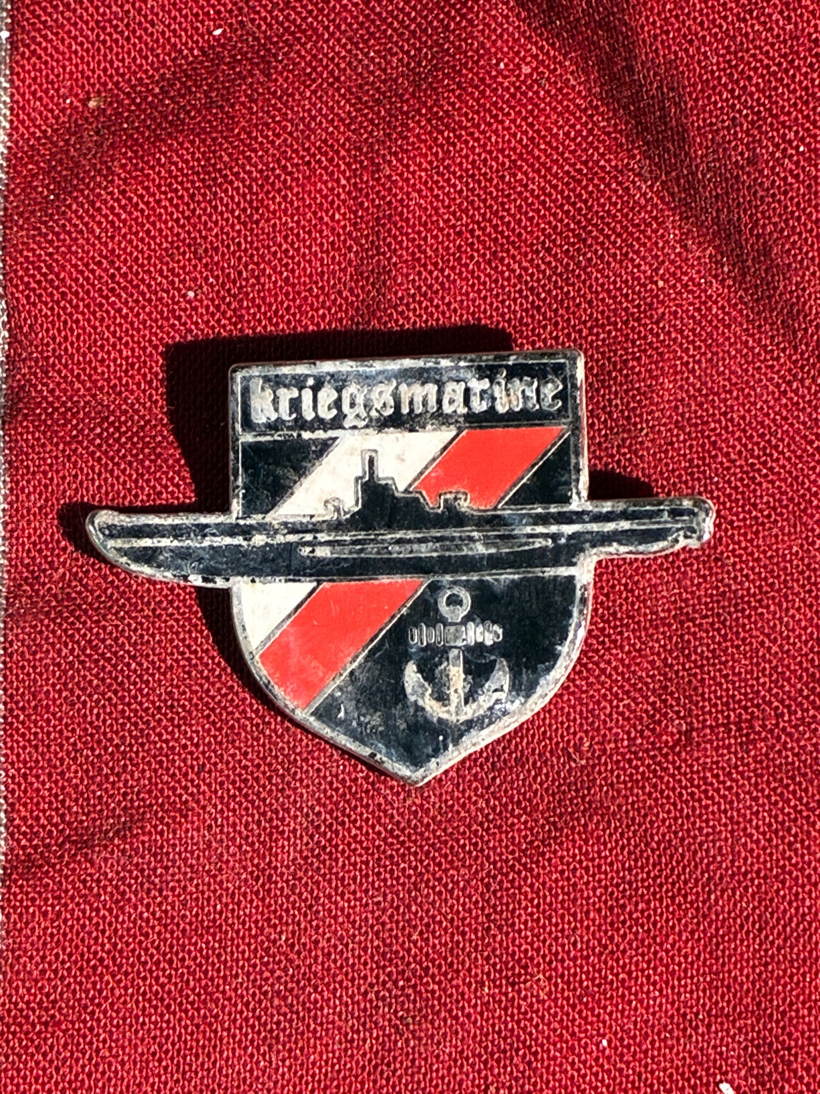 WW2 German Kriegsmarine U-Boat Commemorative Pin (Unofficial): This small enameled pin depicts a stylized German U-boat set against a shield bearing red, white, and black diagonal fields. The top bar features the word “Kriegsmarine”—the navy of Nazi Ger