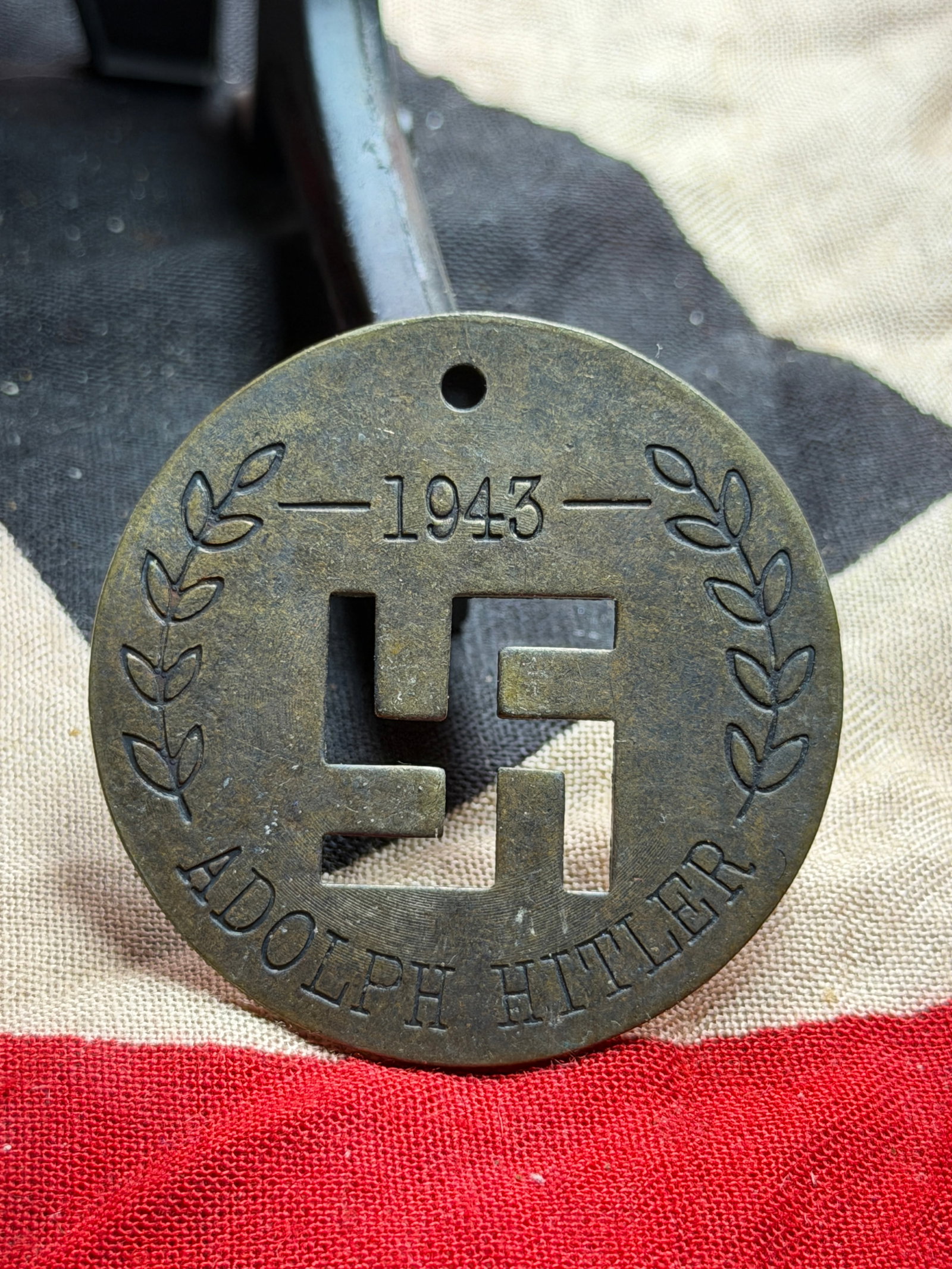 WW2 German Commemorative Medallion for Adolf Hitler, 1943: This circular metal medallion, dated 1943, features a pierced swastika at its center, flanked by laurel branches—classical symbols of victory and authority—encircling the inscription “ADOLPH HIT