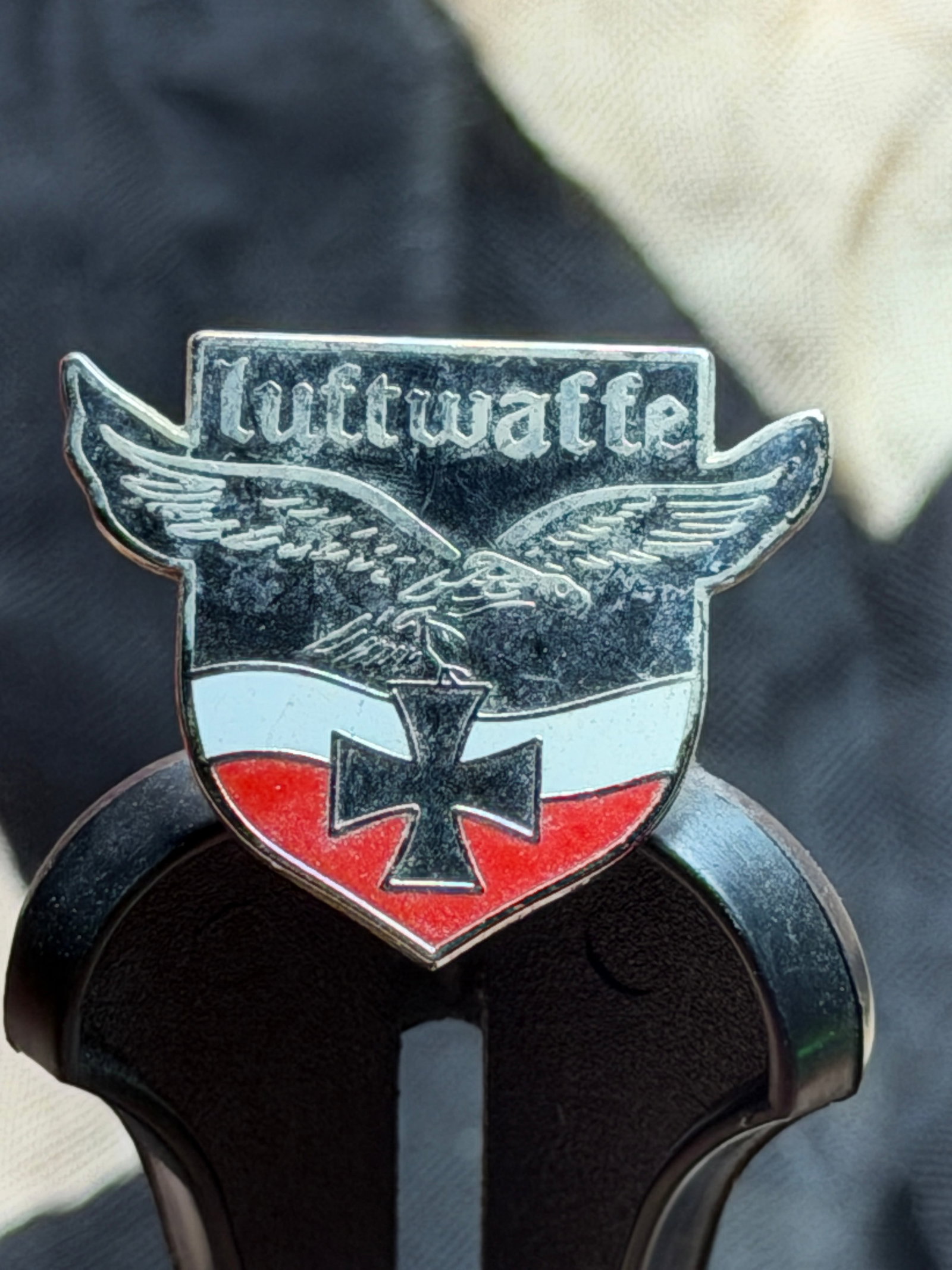 WW2 German Luftwaffe Shield Badge with Iron Cross Motif: This small shield-shaped badge features the inscription “Luftwaffe” above a stylized eagle in flight, with an Iron Cross prominently displayed at the center over a black-white-red field. Produ