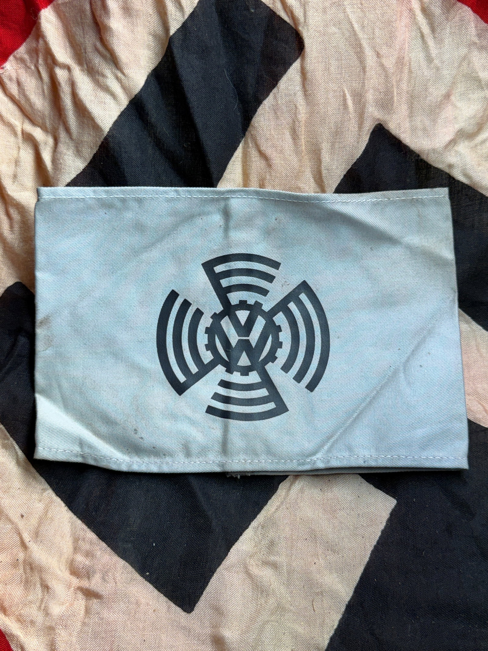 WW2 German Volkswagen Factory Organization Armband (KDF/DAF): This light-gray cotton armband bears the printed emblem of the Volkswagen factory organization—an industrial cog encircling the “VW” monogram, flanked by radiating bands symbolizing mechaniz
