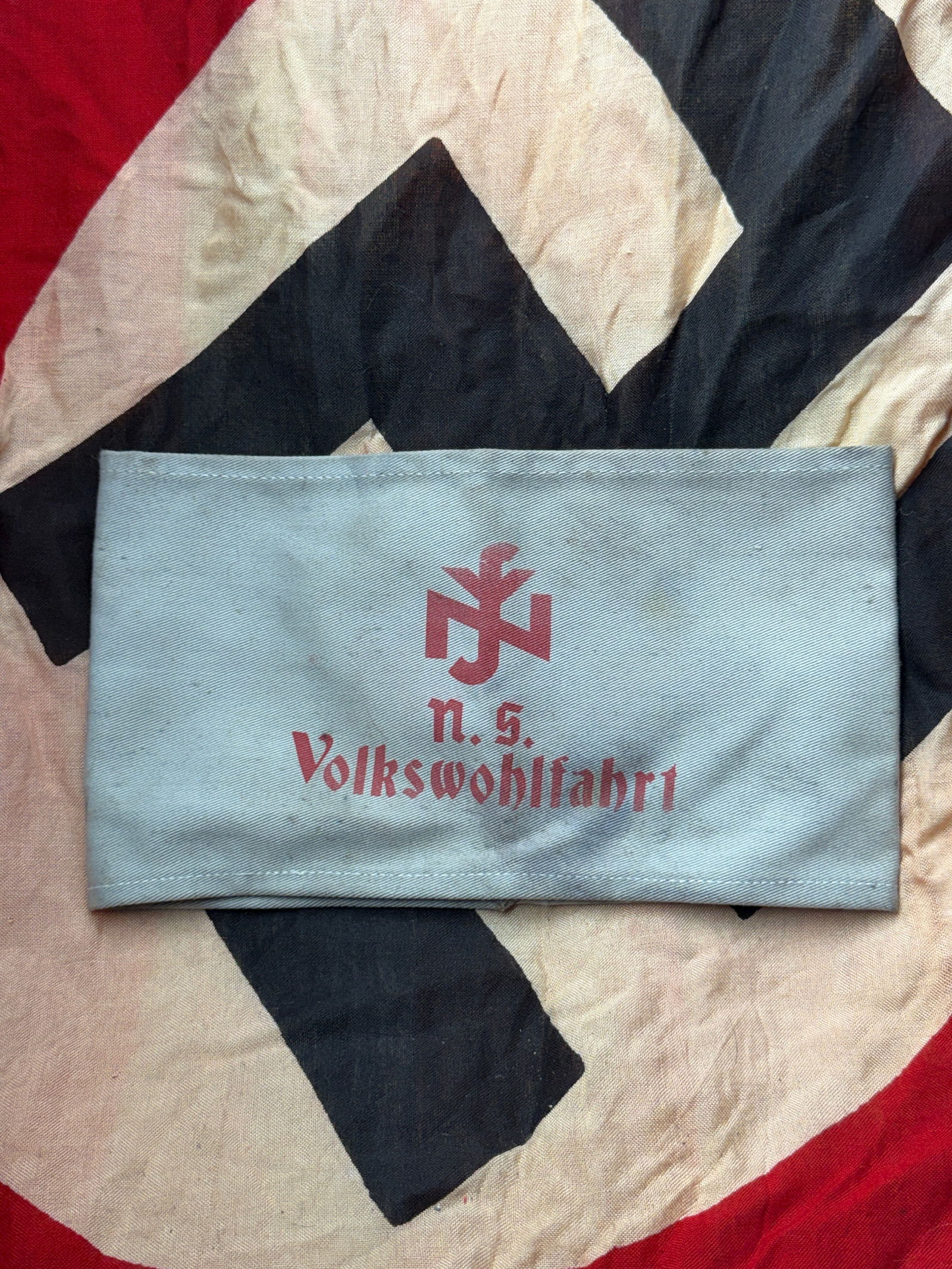 WW2 German NSV (National Socialist People’s Welfare) Armband, Germany, c. 1933–1945: This armband was worn by members or auxiliaries of the Nationalsozialistische Volkswohlfahrt (NSV), the welfare organization of the Nazi Party. Constructed from light grey woven fabric, it bears the r