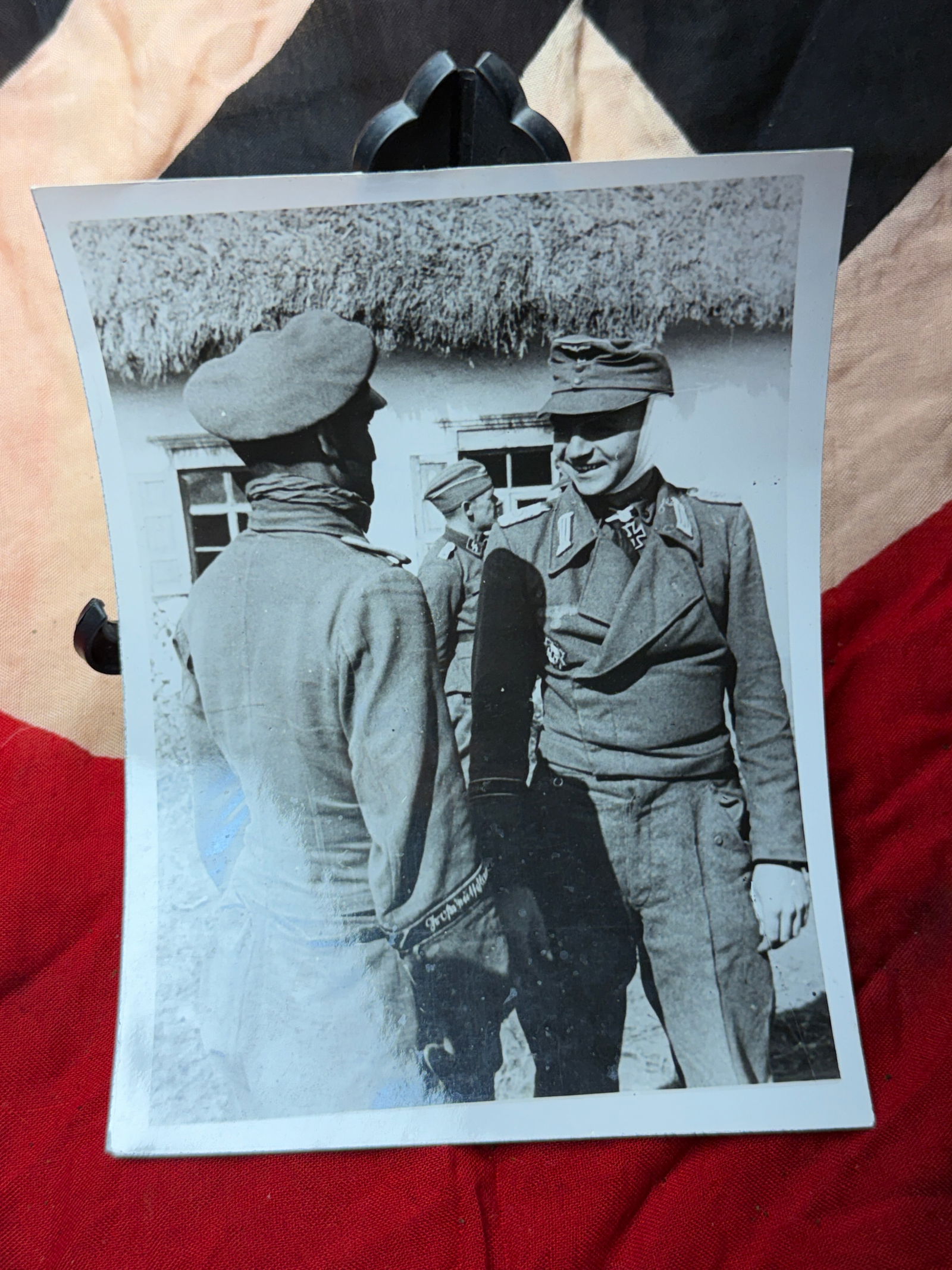 WW2 German Front-Line Officers in Conversation, Eastern Front With Certification: This original wartime photograph depicts German Army officers in informal discussion near a rural structure on the Eastern Front during the Second World War. The figures wear field uniforms typical of