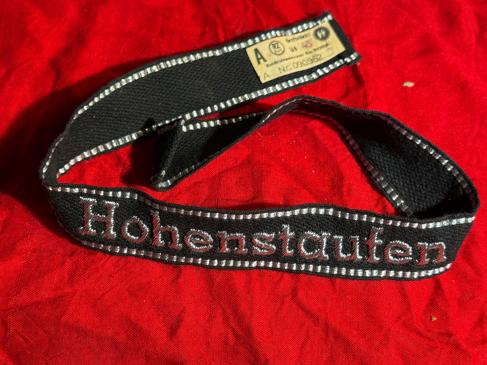 WW2 German Waffen-SS Cuff Title With RZM Paper Tag “Hohenstaufen,?? 9th SS Panzer Division, Nazi: Black cloth Waffen-SS cuff title embroidered with the name “Hohenstaufen”, worn on the uniform sleeve by members of the 9th SS Panzer Division “Hohenstaufen.” The band features silver-
