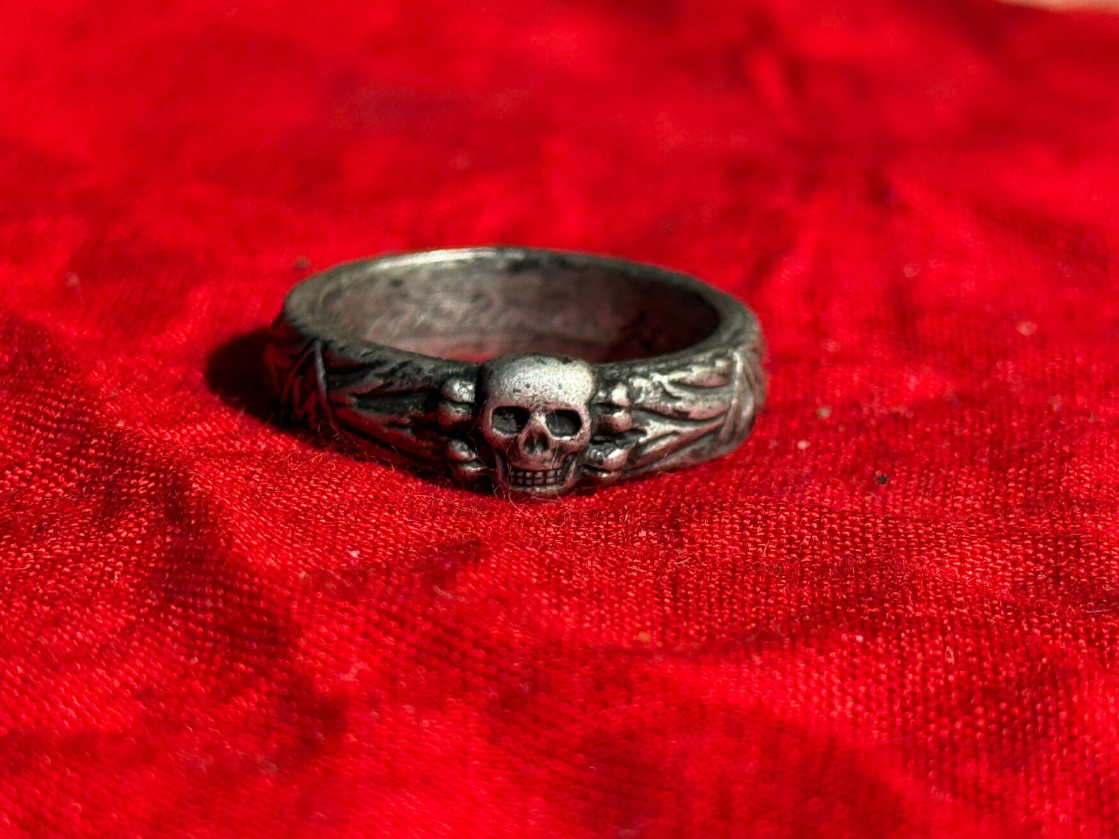 WW2 German Totenkopf Motif Ring (Patriotic or Soldier’s Souvenir Ring): Metal ring featuring a Totenkopf (death’s head) skull motif with crossed bones, flanked by decorative symbols including a swastika within a circle and geometric or runic-style designs along the band