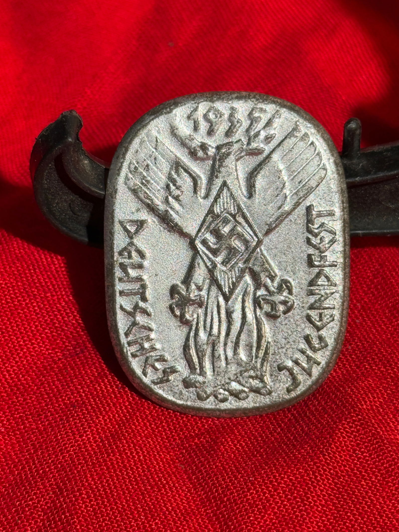 WW2 German Hitler Youth “Adolf Hitler – Dank der Nation?? Commemorative Tinnie Badge, 1937: Small stamped metal commemorative badge featuring the Hitler Youth (Hitlerjugend) diamond emblem with swastika, flanked by stylized wings and above a flaming torch. The inscription reads “Adolf Hitl