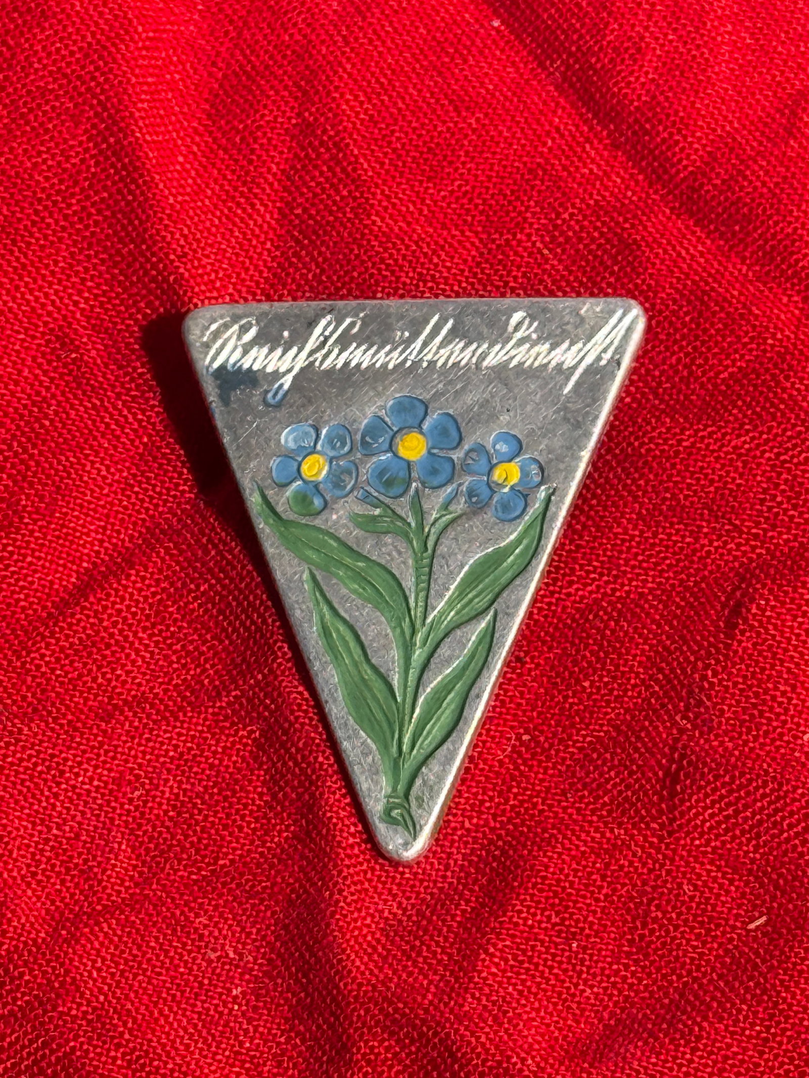 WW2 German Winterhilfswerk “Vergissmeinnicht?? Reichsmütterdienst Donation Badge: Triangular painted aluminum badge depicting a cluster of blue forget-me-not flowers beneath the inscription “Vergissmeinnicht.” Produced by Fries-Beuster & Oehlschlägel of Berlin-Hohenschönh