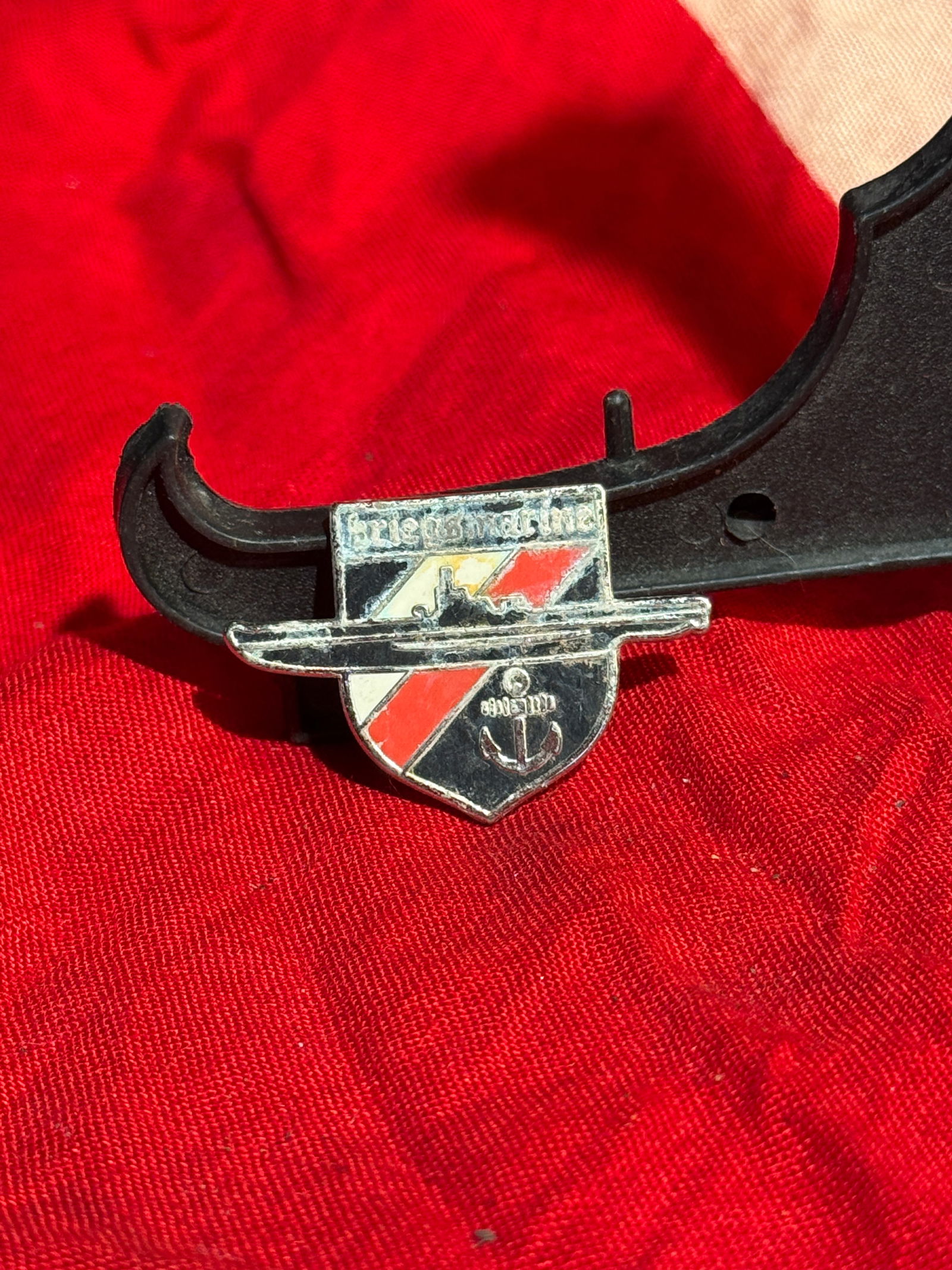 WW2 German Kriegsmarine Commemorative Lapel Badge: Small enamel Kriegsmarine-themed lapel badge featuring a shield with black, white, and red diagonal stripes, a naval anchor, and a warship silhouette. The top inscription appears to reference “Krieg