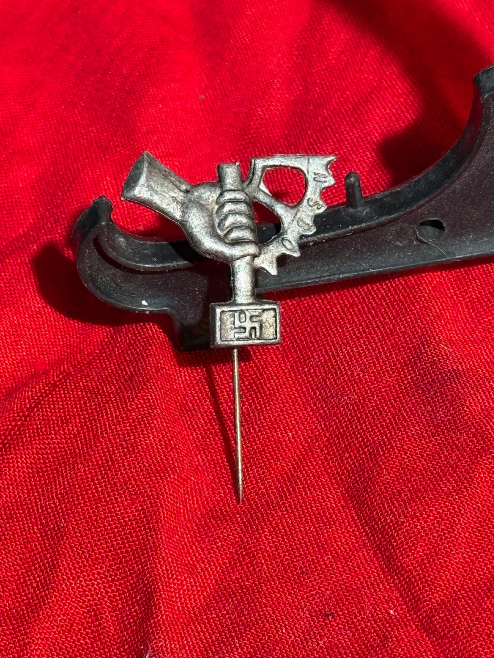 WW2 German Labor-Themed Lapel Stick Pin, 1930s–1940s: Small metal lapel stick pin depicting a clenched hand holding a hammer beside a gear wheel, symbols associated with industrial labor and technical trades. A small plaque below bears a swastika emblem,