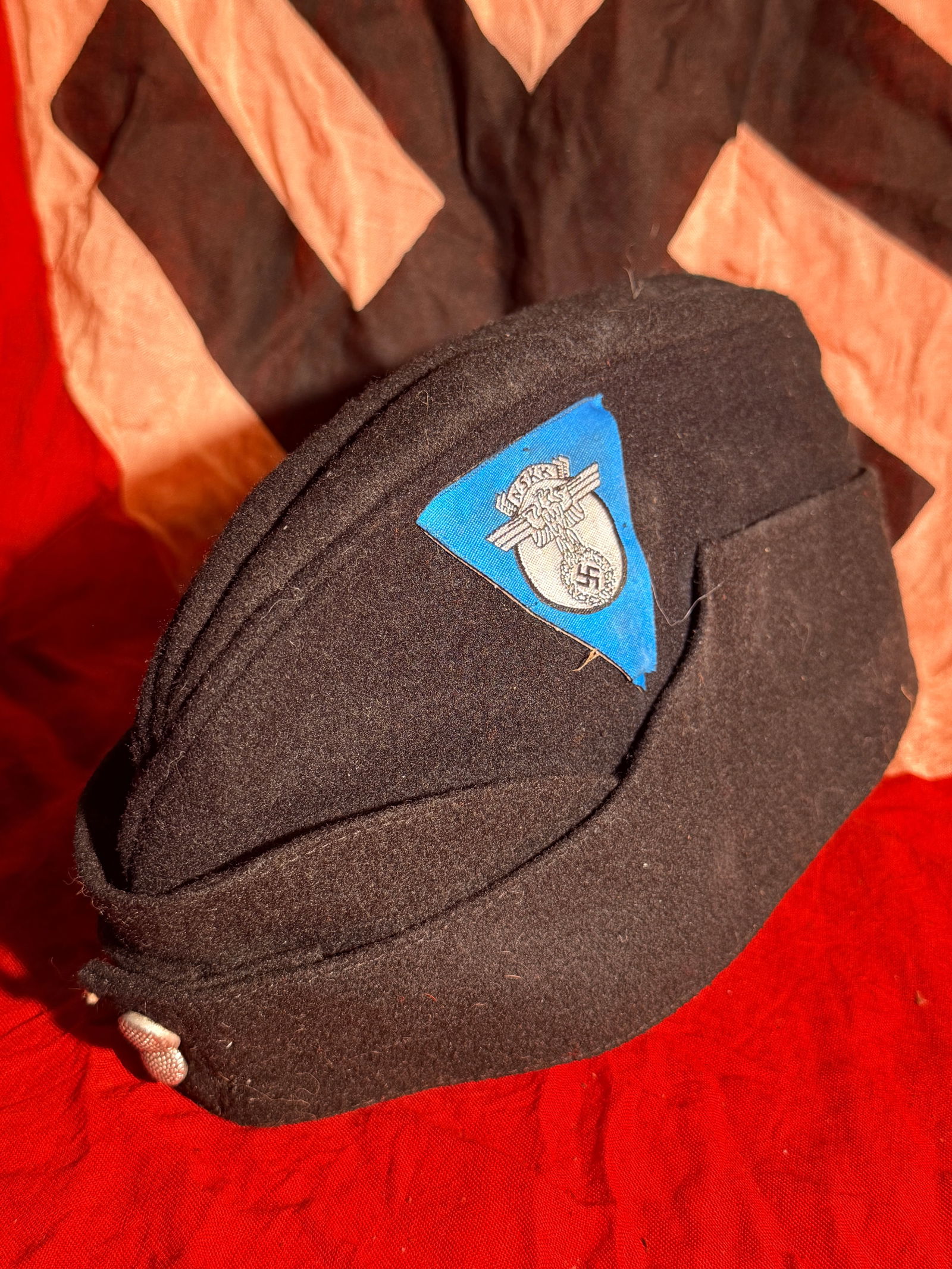 WW2 German NSKK Side Cap (Schiffchen), Germany, c. 1930s With RZM Paper Tag: Black wool side cap (Schiffchen) worn by members of the National Socialist Motor Corps (NSKK) during the Third Reich period. The cap features a triangular blue NSKK insignia patch displaying the organ
