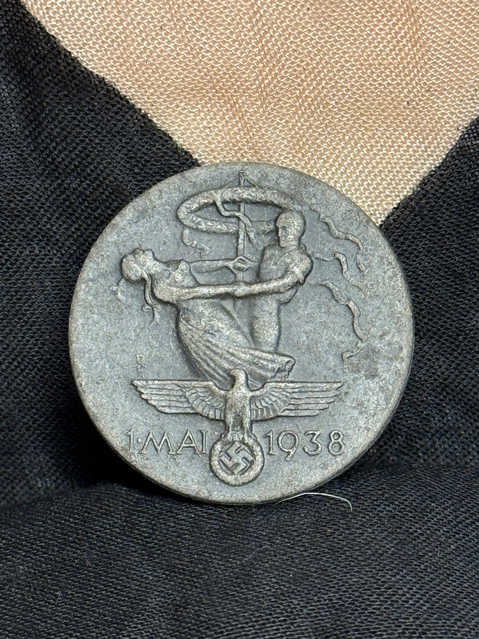 WW2 German “1 May 1938?? Commemorative Tinnie (Day of National Labour Badge): A stamped metal badge commemorating May Day (Tag der Arbeit) celebrations in 1938. The design shows a male and female figure symbolizing German labor and unity, above a national eagle and date inscrip