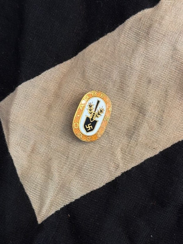 WWII German Agricultural or Organizational Membership Badge (“Arbeits?? Inscription): This oval enamel badge features a stylized agricultural motif—tools and grain—above a swastika, surrounded by the word “Arbeits” (labor/work). Badges of this type were associated with labo