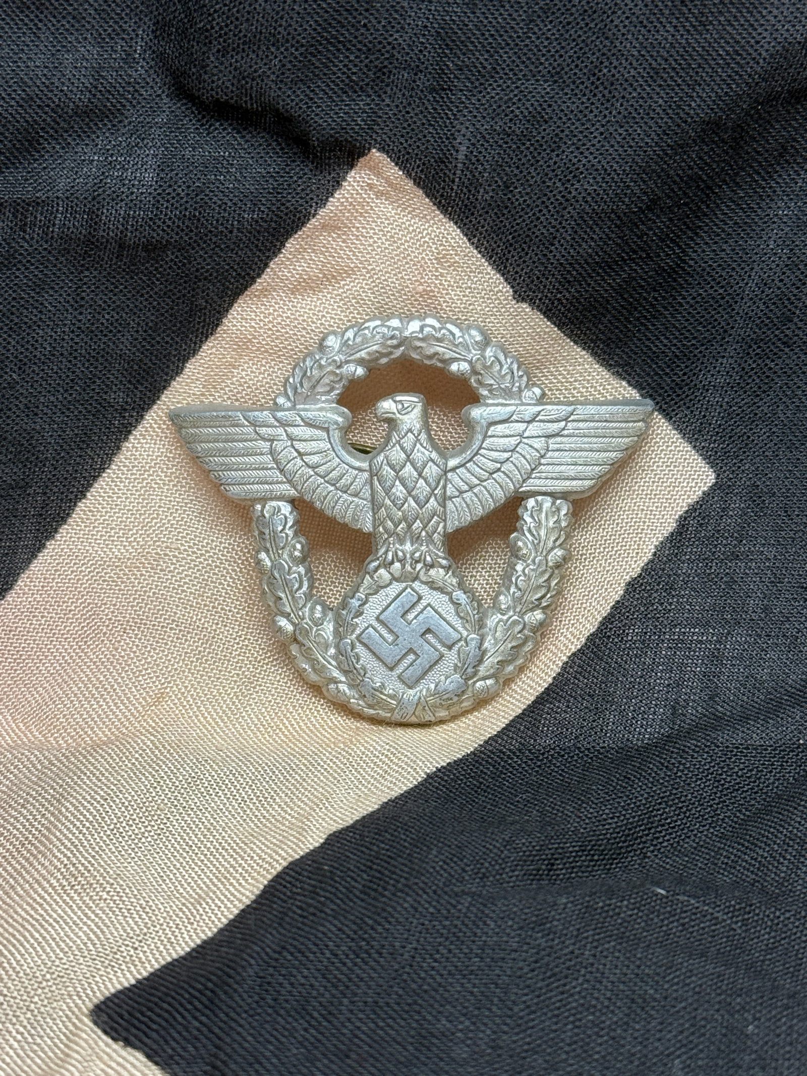WWII German Police/State Service Eagle Wreath Cap Badge: This aluminum cap badge depicts a spread-wing eagle clutching a swastika within an oak-leaf wreath—an emblem commonly associated with German police or state service organizations during WWII. The wr