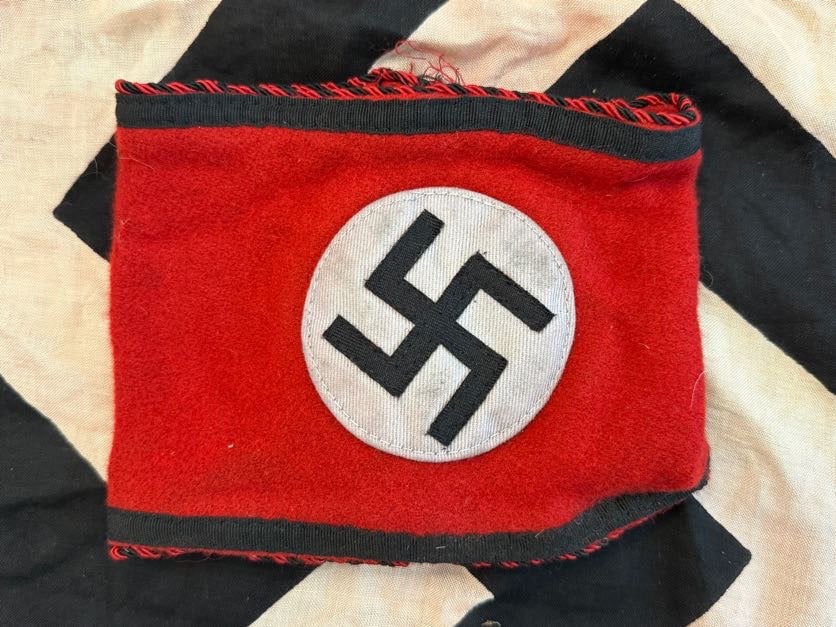 WWII German SS Armband with RZM Tag: This red armband features a central white roundel with a black swastika, a widely recognized emblem of Nazi Germany. The piece is trimmed with black and red twisted cord edging, suggesting a higher-qu