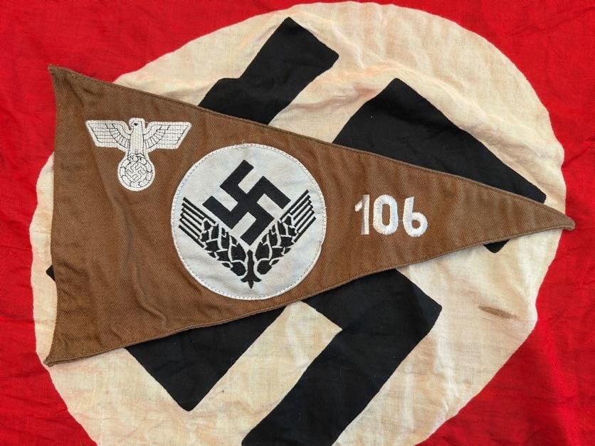 WWII German NSDAP/SA Pennant with Eagle and Swastika: This triangular pennant features a brown field with a white roundel containing a black swastika, flanked by stylized wings and accompanied by a German eagle emblem. The number “106” is printed