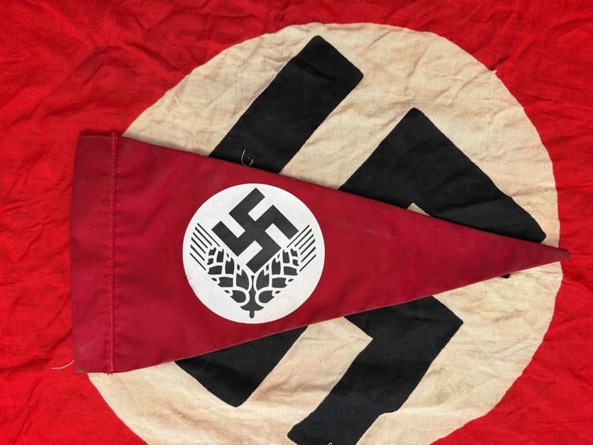 WWII German RAD (Reichsarbeitsdienst) Pennant with Wheat Emblem: This triangular pennant features a red field with a white roundel containing a black swastika above stylized wheat sheaves, a design associated with the Reichsarbeitsdienst (RAD), the state labor serv