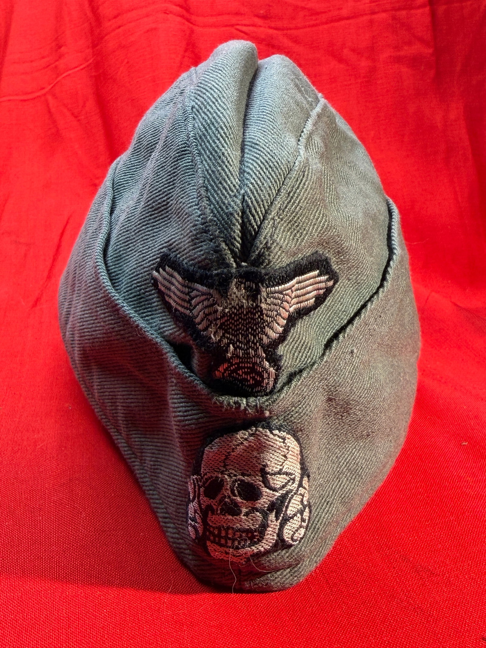 WW2 German SS Totenkopf Overseas Side Cap (Schiffchen) with Eagle and Skull Insignia: WW2-era German SS overseas side cap (Schiffchen style) featuring the distinctive Totenkopf (skull) insignia beneath a national eagle emblem on the front. The cap is constructed in field-gray twill fab