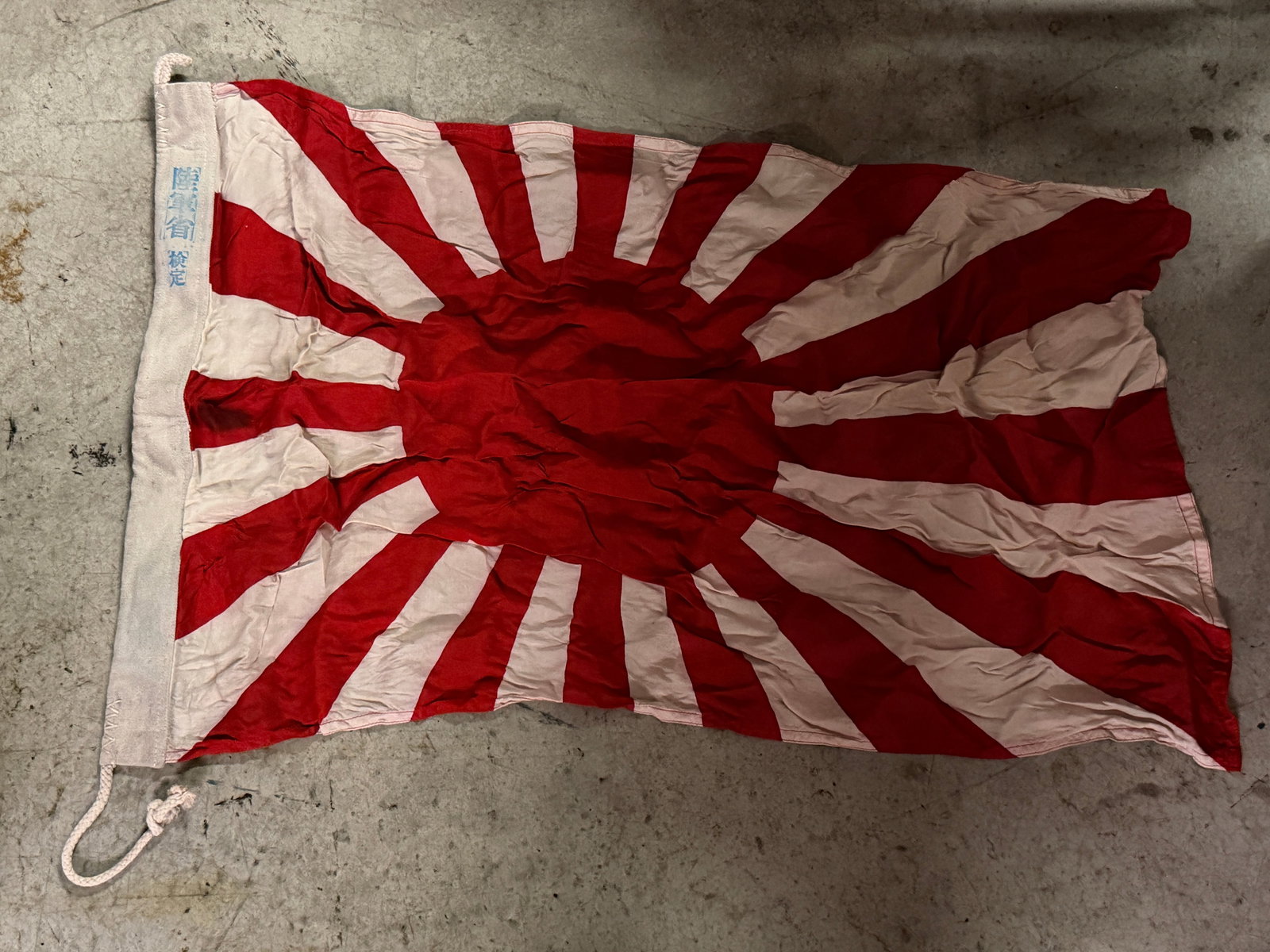 WWII Japanese Rising Sun Flag with Canvas Hoist and Markings: This red and white Rising Sun flag features a bold radiating sunburst design extending outward from a central disc, rendered on a lightweight fabric that shows visible creasing and wear from handling