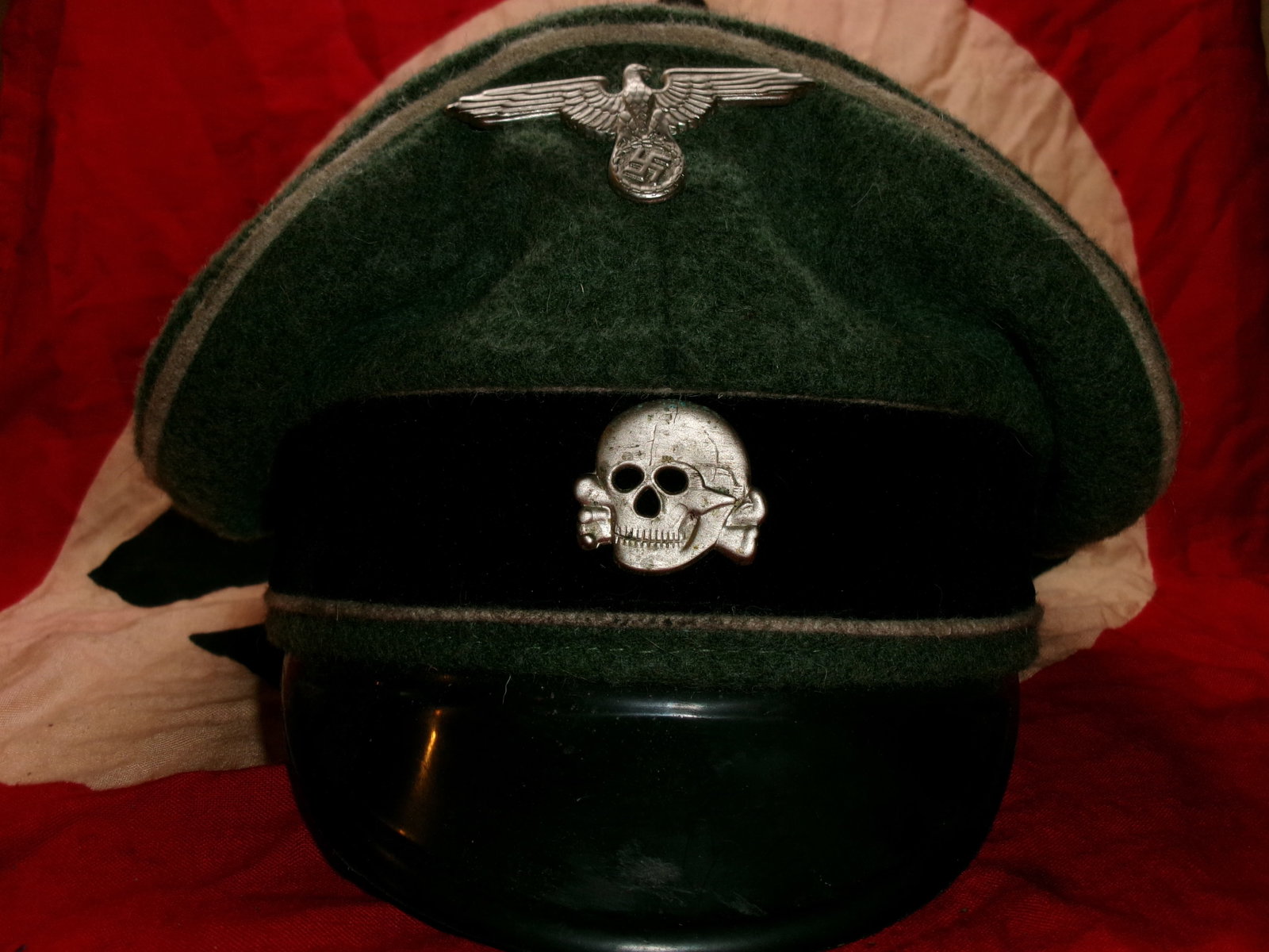 WWII German SS Officer’s Visor Cap (Totenkopf Insignia): A dark wool visor cap bearing the skull-and-crossbones (Totenkopf) emblem, associated with the SS (Schutzstaffel). The additional eagle insignia above the skull denotes rank and affiliation within Naz