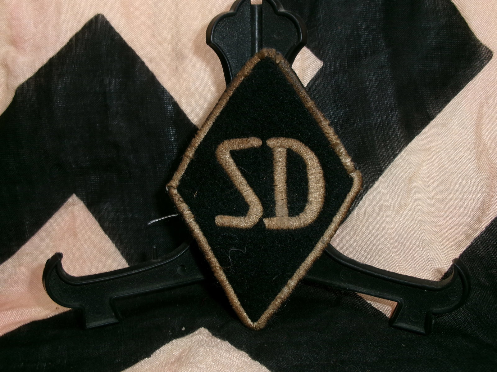 WWII German Sicherheitsdienst (SD) Sleeve Diamond Insignia: This black cloth diamond-shaped patch bearing the white “SD” letters represents the Sicherheitsdienst (Security Service), the intelligence agency of the SS during World War II. Worn on the low