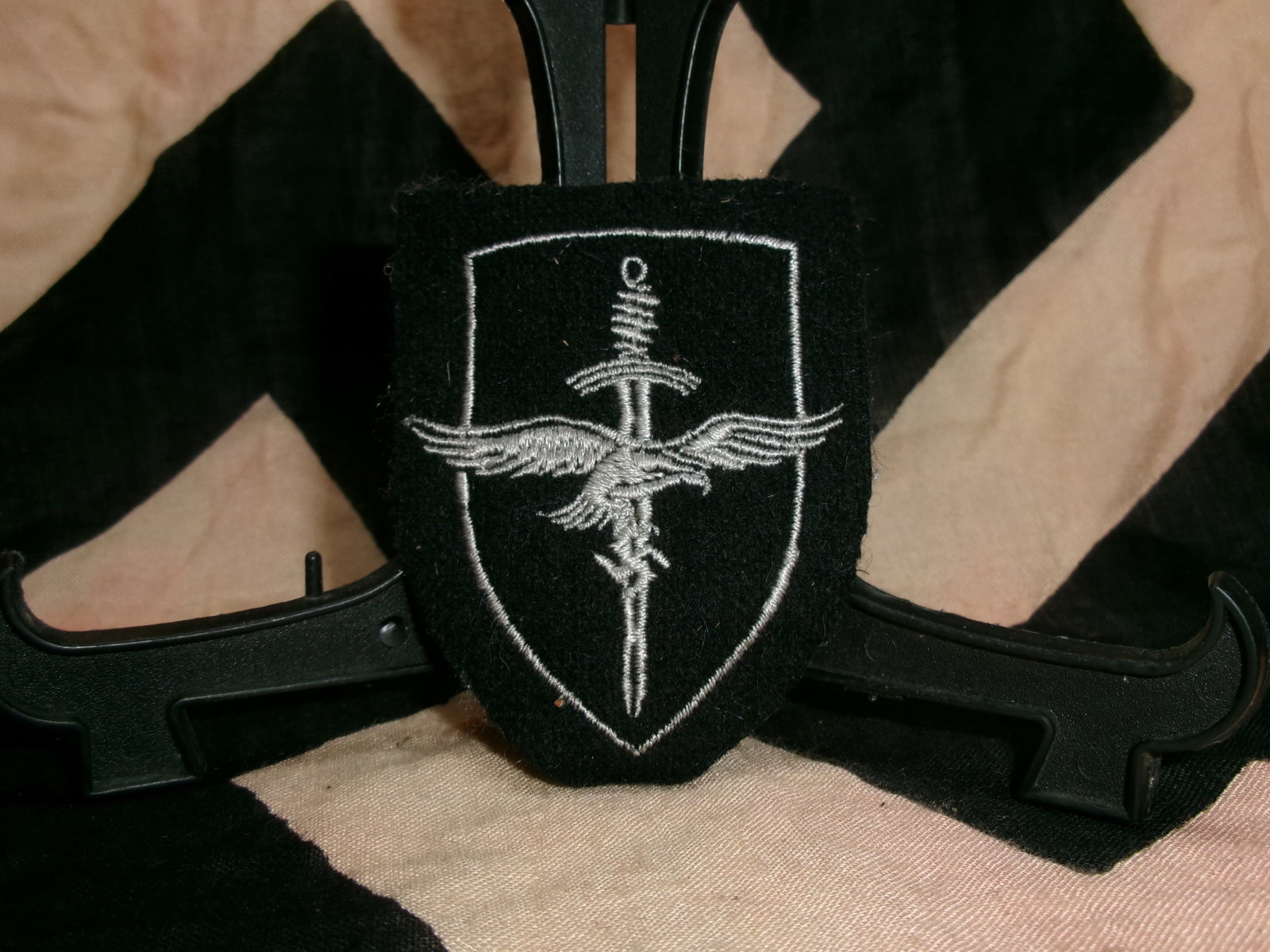 WWII German Luftwaffe womens ‘Helferinnen’ sleeve patch: This black cloth sleeve patch features a striking white embroidered design consisting of a vertical sword overlaid with outstretched wings, all enclosed within a shield-shaped border. The imagery comb