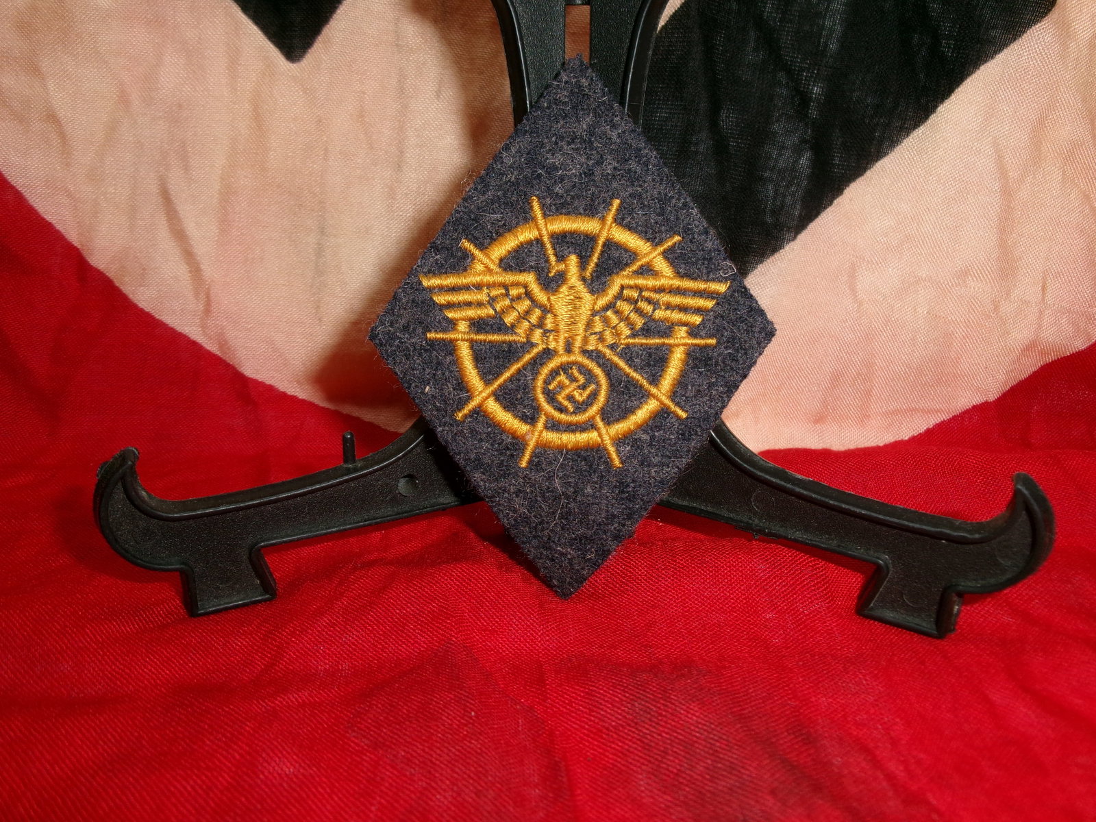WWII German Signals/Communications-Style Sleeve Insignia (Winged Transmission Motif): This diamond-shaped cloth insignia features intricate gold embroidery depicting a winged central device surrounded by a circular framework with radiating lines resembling lightning bolts or transmissi