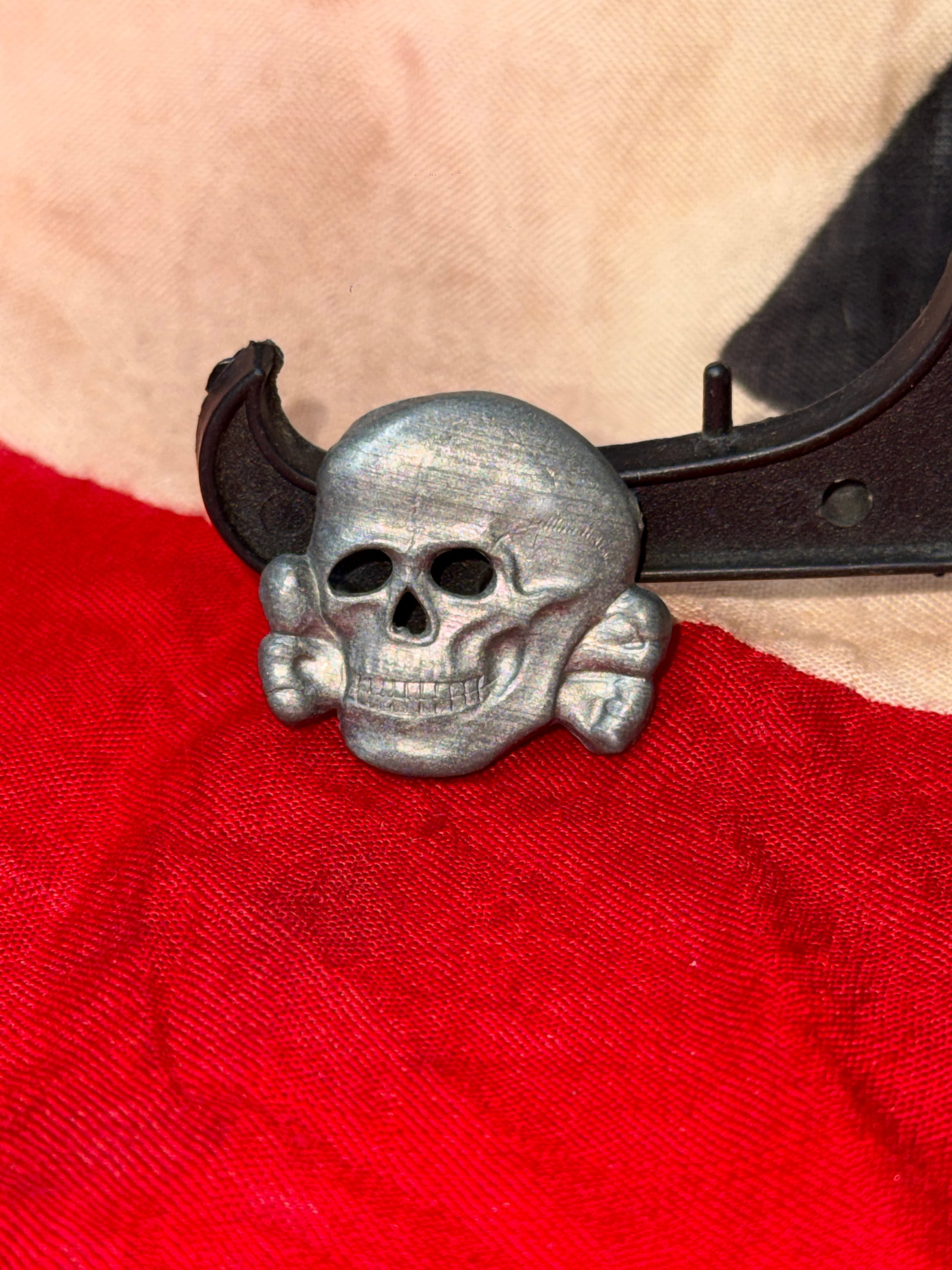 WWII German Skull & Crossbones Metal Cap Badge (Totenkopf Style): This metal cap badge features a skull and crossbones design commonly referred to as the “Totenkopf.” The piece has a matte silver-toned finish with detailed facial features, hollow eye sockets