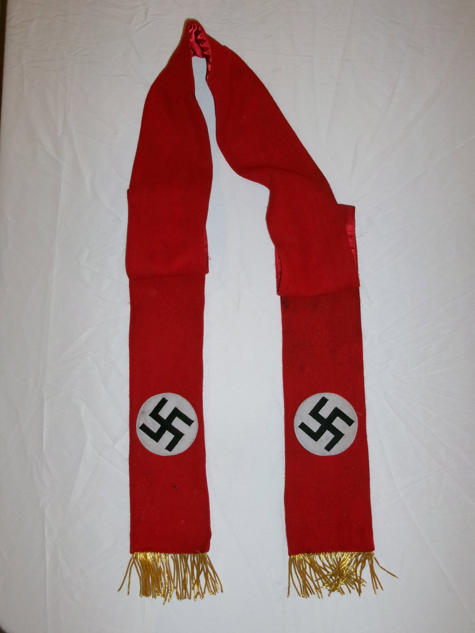 WWII German Political Organization Ceremonial Scarf (Red with Swastika Roundels): This red cloth scarf features two circular white disks bearing black swastika emblems, along with gold-colored fringe at each end. The design and coloration are consistent with ceremonial or political
