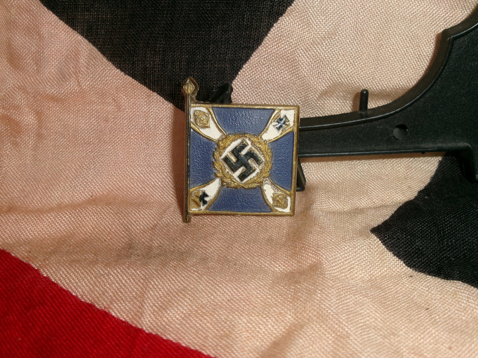 WWII German Organization Flag Lapel Pin (Enamel Swastika Emblem): A small enamel lapel pin designed in the form of a stylized flag bearing a central swastika within a wreath. Such pins were worn by civilians or party affiliates to signify allegiance to National Soci
