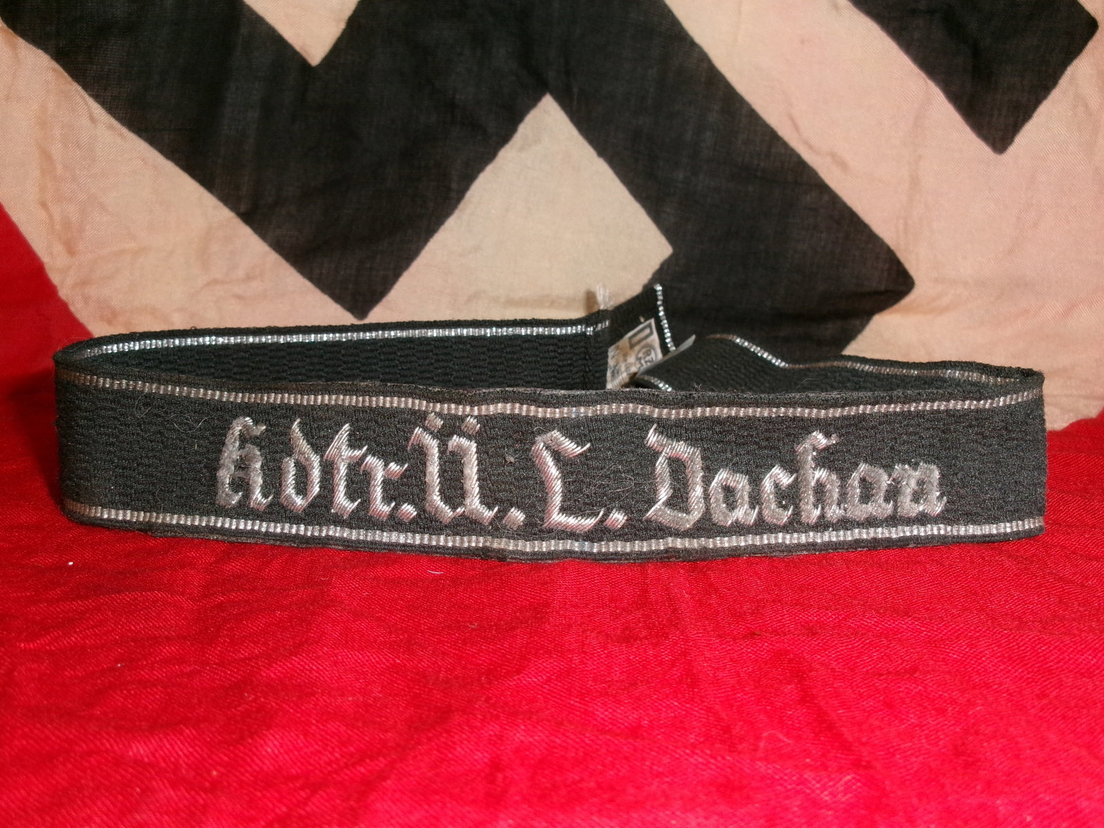 WWII German SS Cuff Title “Kdtr. II./L. Dachau?? – RZM Tagged: Black woven cuff title featuring the silver script “Kdtr. II./L. Dachau”, associated with SS personnel and Dachau command structure. The band exhibits BeVo-style construction with silver borde