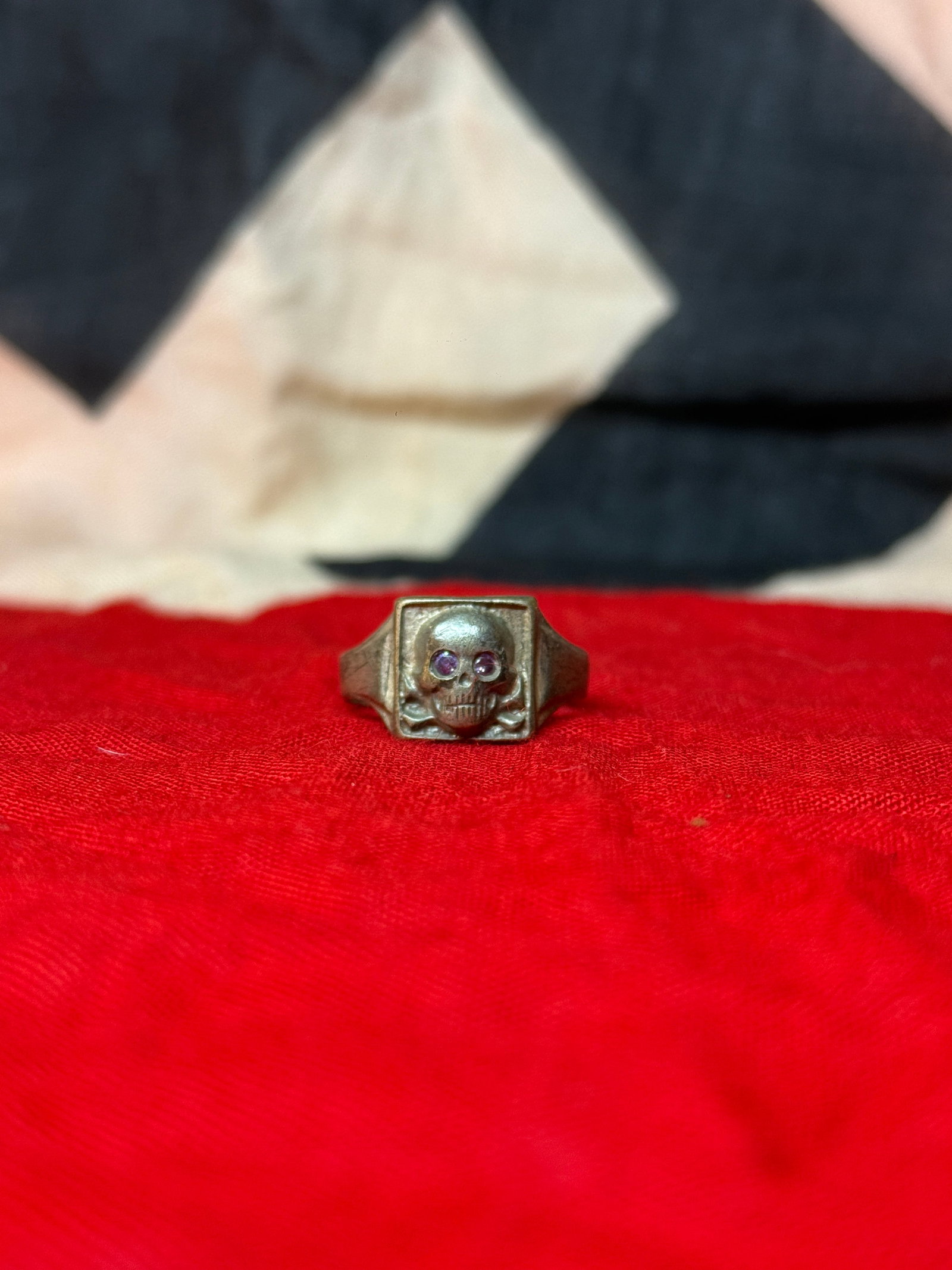 WWII German Totenkopf Skull Ring with Stone-Set Amethyst Stone Eyes: This metal ring features a prominent Totenkopf (death’s head) motif, with a skull and crossbones rendered in relief on a square bezel. The eye sockets are set with small colored stones, adding a dis