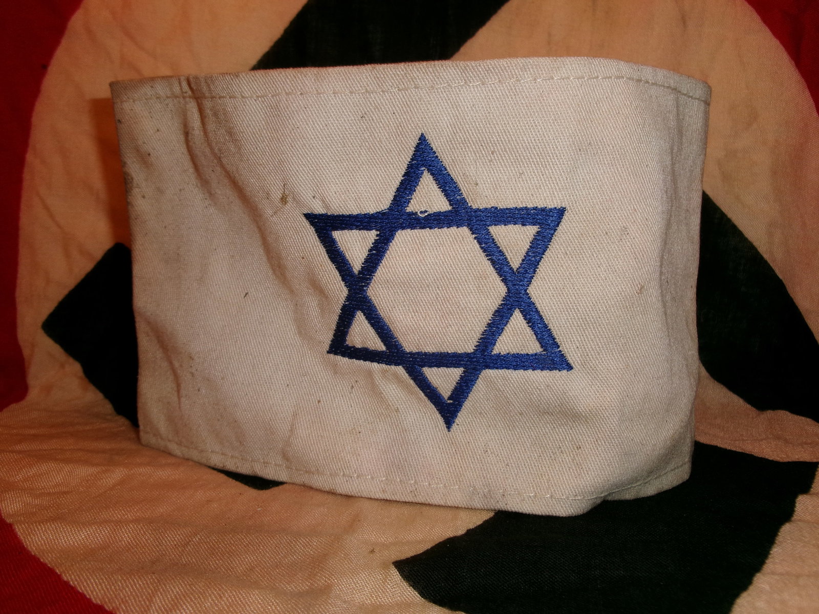 WWII German Jewish Identification Armband With Star Of David: This white cloth armband bears a prominently embroidered blue Star of David, a symbol imposed under wartime regulations to visibly identify Jewish individuals within territories controlled by Nazi Ger