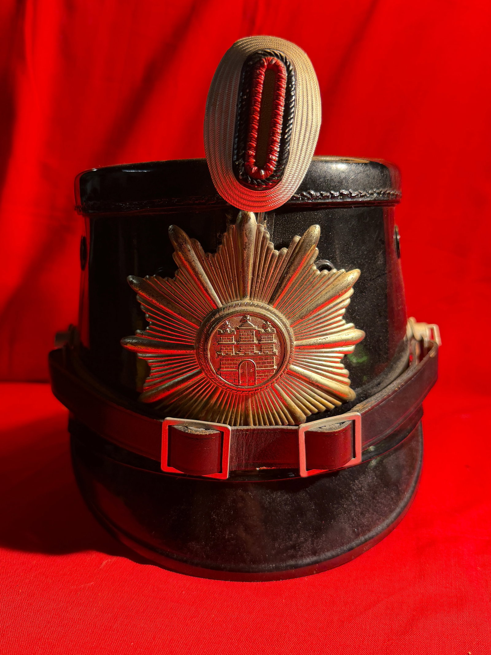 WWII German Police / Civic Visor Helmet (Shako Style) – By Robert Lubstein: Black lacquered visor-style helmet featuring a prominent multi-piece metal starburst plate with central city-style crest, topped with a ribbed cockade displaying a red center. The front retains its or