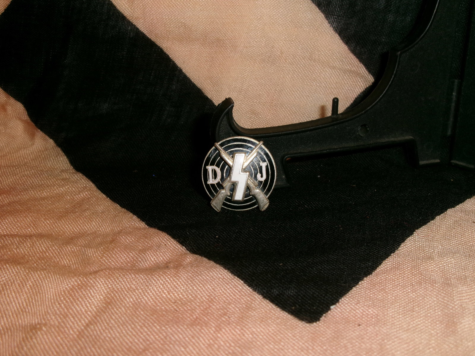 WWII German DJ (Deutsches Jungvolk) Shooting Proficiency Badge By RZM M1/123: This circular badge features the intertwined initials “DJ” flanking a central sig rune above crossed rifles, identifying it as an award connected to the Deutsches Jungvolk, the junior branch o