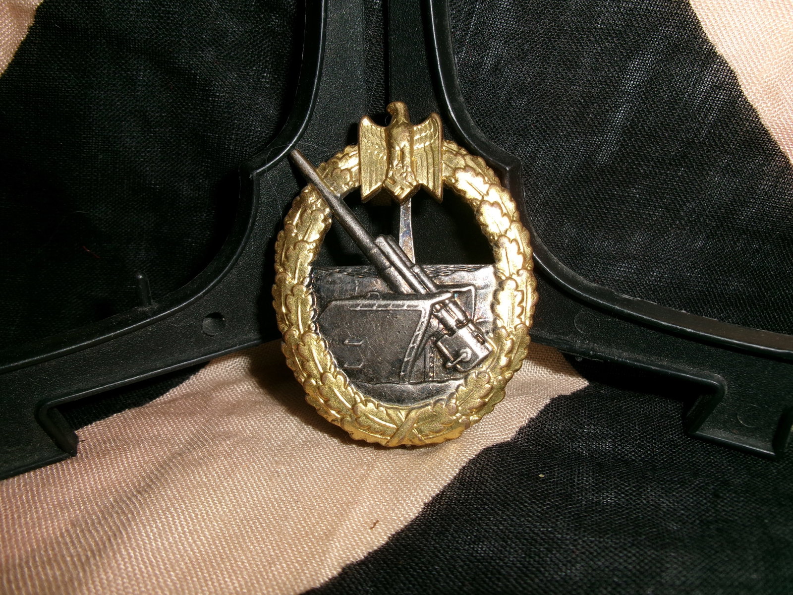 WWII German Kriegsmarine Coastal Artillery War Badge (Kriegsabzeichen für die Marineartillerie) By: This Kriegsmarine Coastal Artillery War Badge features a gilt oakleaf wreath encircling a coastal gun emplacement with a rising barrel, surmounted by the national eagle clutching a swastika. Institute