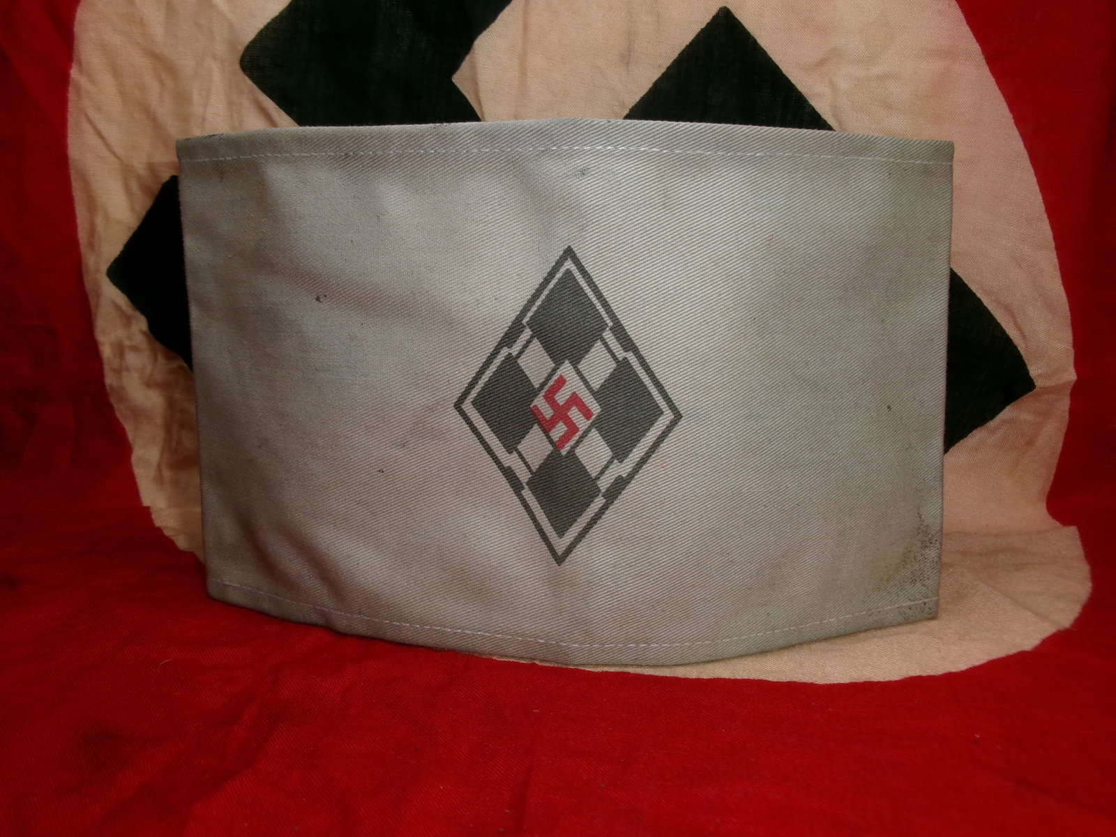 WWII Hitler Youth Armband (HJ Armband): This red cloth armband displays a central white lozenge with a black swastika, associated with the Hitler Youth organization. The interior shows manufacturing marks and wear consistent with use. Armba