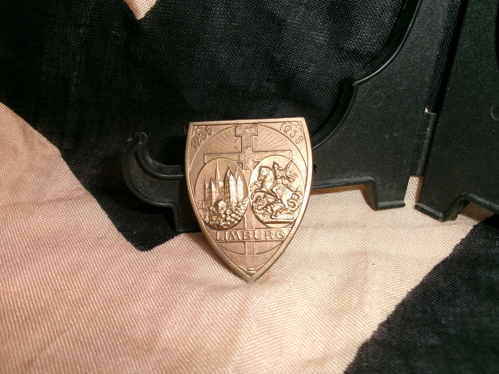 WWII German Limburg Commemorative Badge (1935 Civic or Regional Issue): A metal badge marked “LIMBURG” and dated 1935, featuring a split design: one side depicting architectural elements of the town and the other a mounted figure, likely St. George, above a styliz