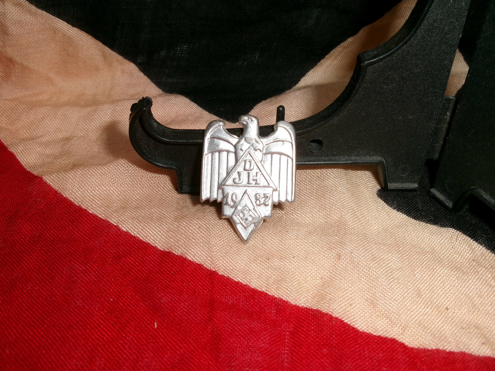 WWII German Hitler Youth Proficiency Badge (Leistungsabzeichen), Eagle over Shield Design: A die-struck Hitler Youth proficiency badge depicting a spread-wing eagle above a triangular shield bearing stylized lettering and the date “1933,” with a diamond-shaped swastika emblem below.